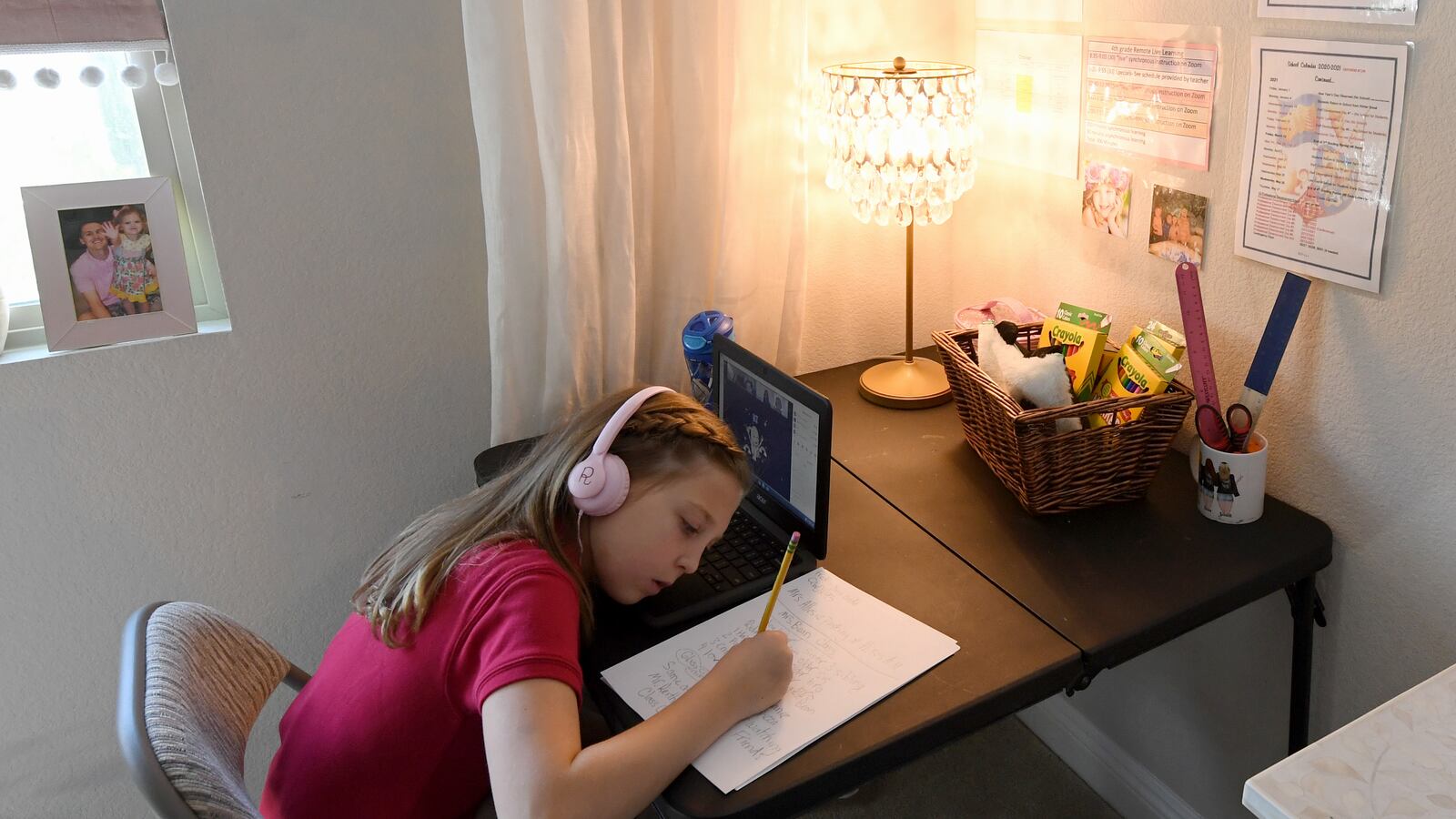 A young girl wearing a pink shirt and headphones writes on a piece of paper while sitting at her desk with a laptop open in front of her.