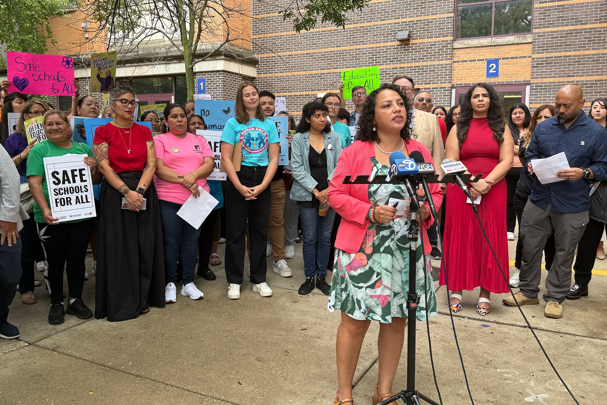 A photograph of a group of people holding signs standing behind a woman speaking behind a group of microphones all in front of a brick school building.