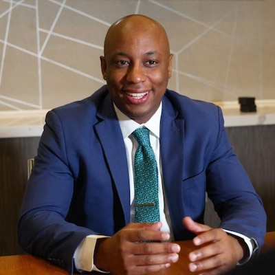 A photograph of a Black man in in a blue suit smiling while talking with his hands.