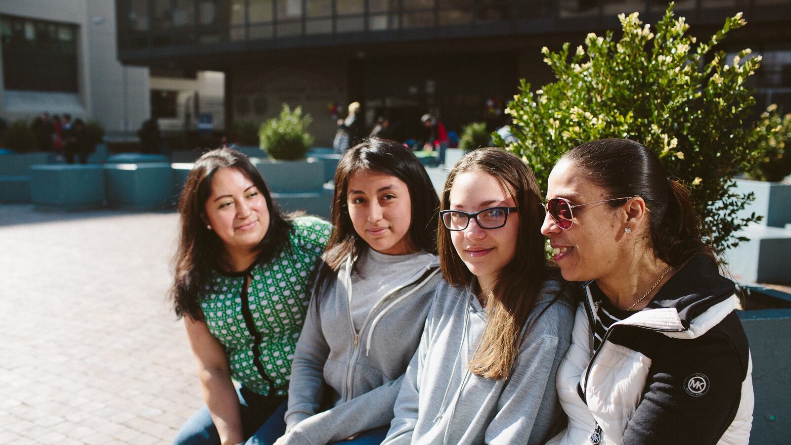 From left, Lordes Lliguichiuzhca, her daughter Elizabeth Cuzco, 13, Sonia Bucur, 13, and her mother Dina Bucur, sit in front of Martin Luther King High School after attending the Round 2 high school fair.