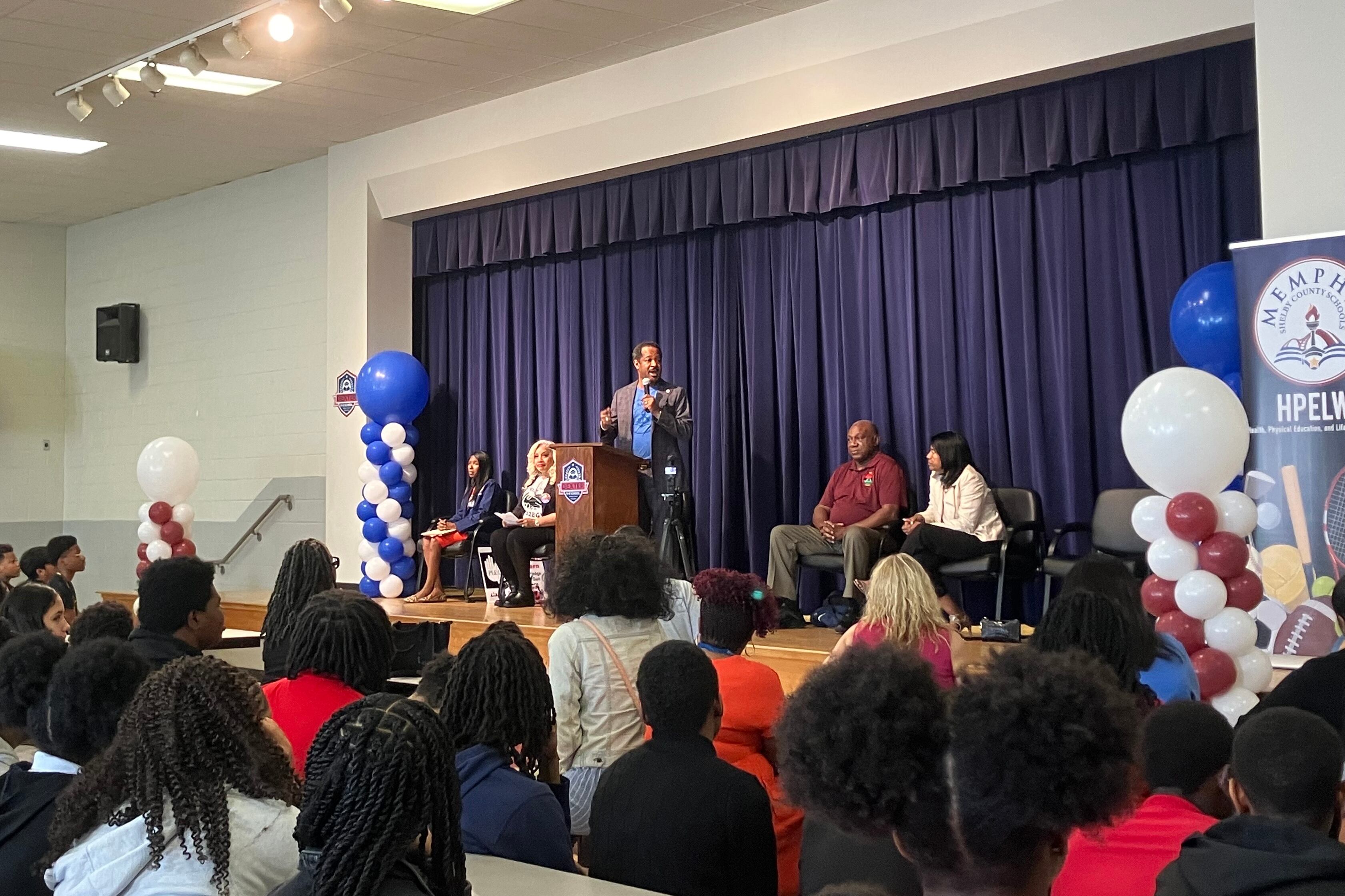 A photograph of a Black man in a suit speaking from a stage in a school building with people sitting on stage and in the audience in chairs.