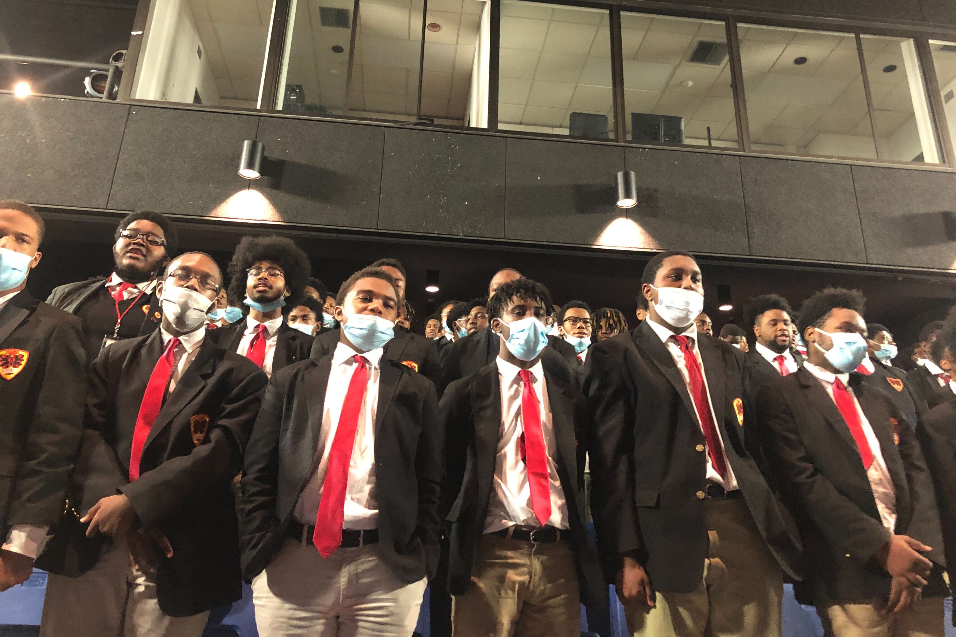 A group of high school students stand at left. Adults are seated nearby in an auditorium.