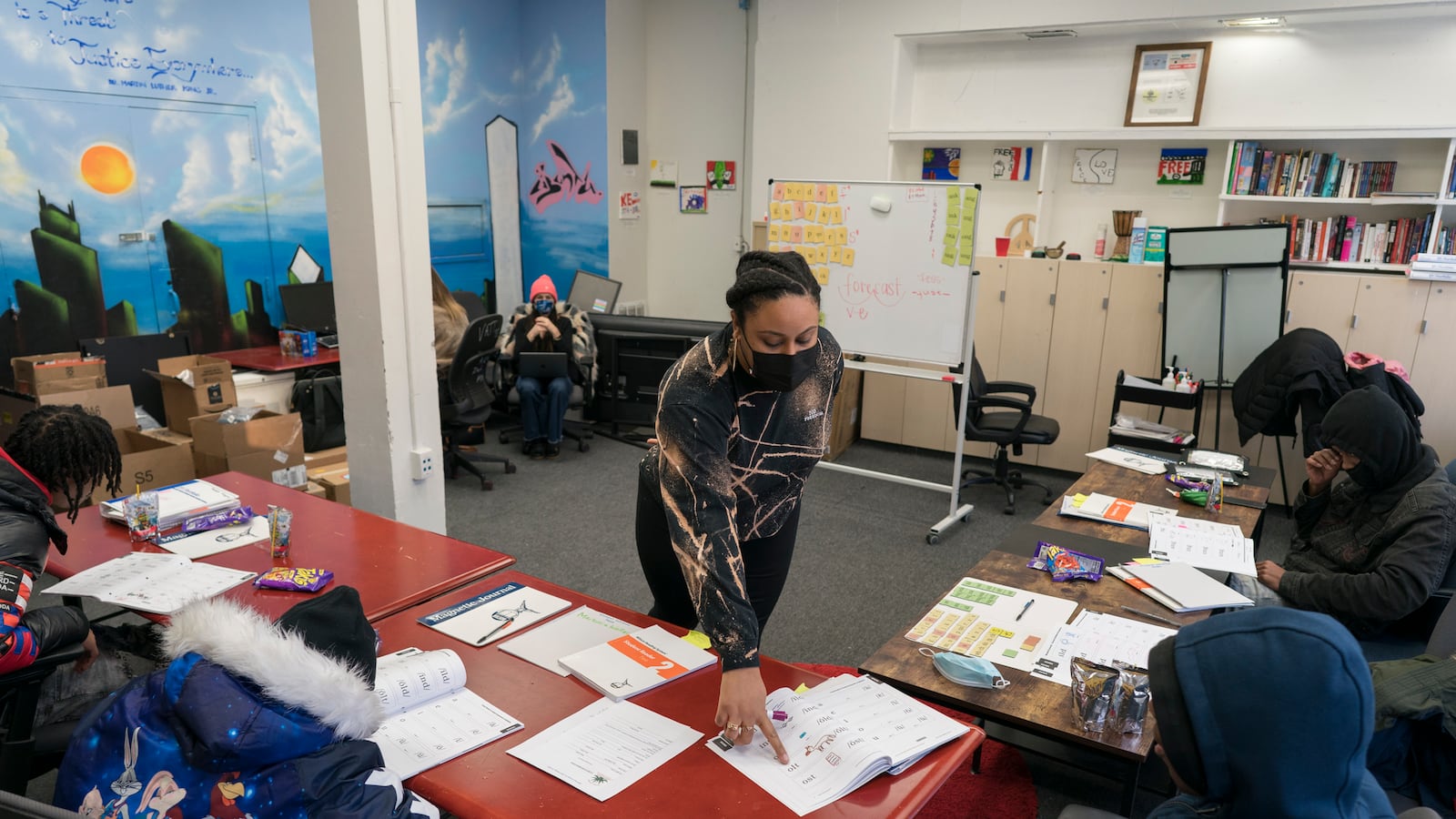 A woman points to a notebook during literacy instruction in a small classroom, as students work on reading skills.