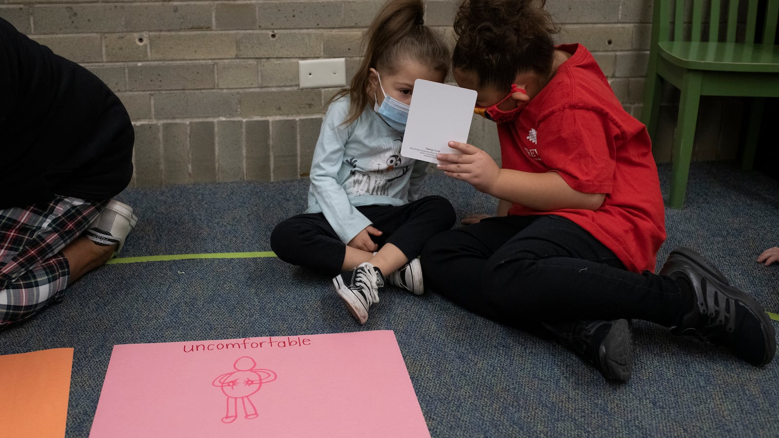 Two young girls hold up a card as they talk during a social-emotional learning lesson. They sit on a grey carpet, and there is a drawing in front of them on pink paper that says “uncomfortable” with a drawing of a person holding their abdomen.