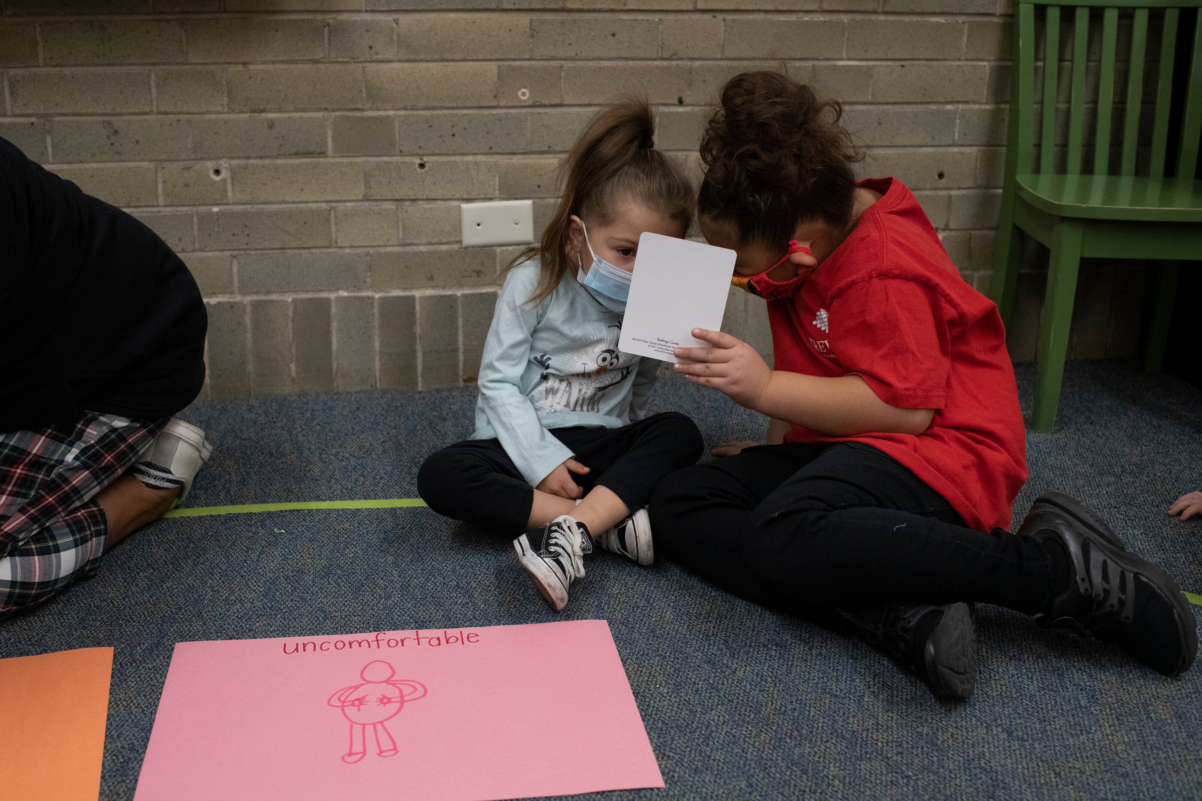 Two young girls hold up a card as they talk during a social-emotional learning lesson. They sit on a grey carpet, and there is a drawing in front of them on pink paper that says “uncomfortable” with a drawing of a person holding their abdomen.