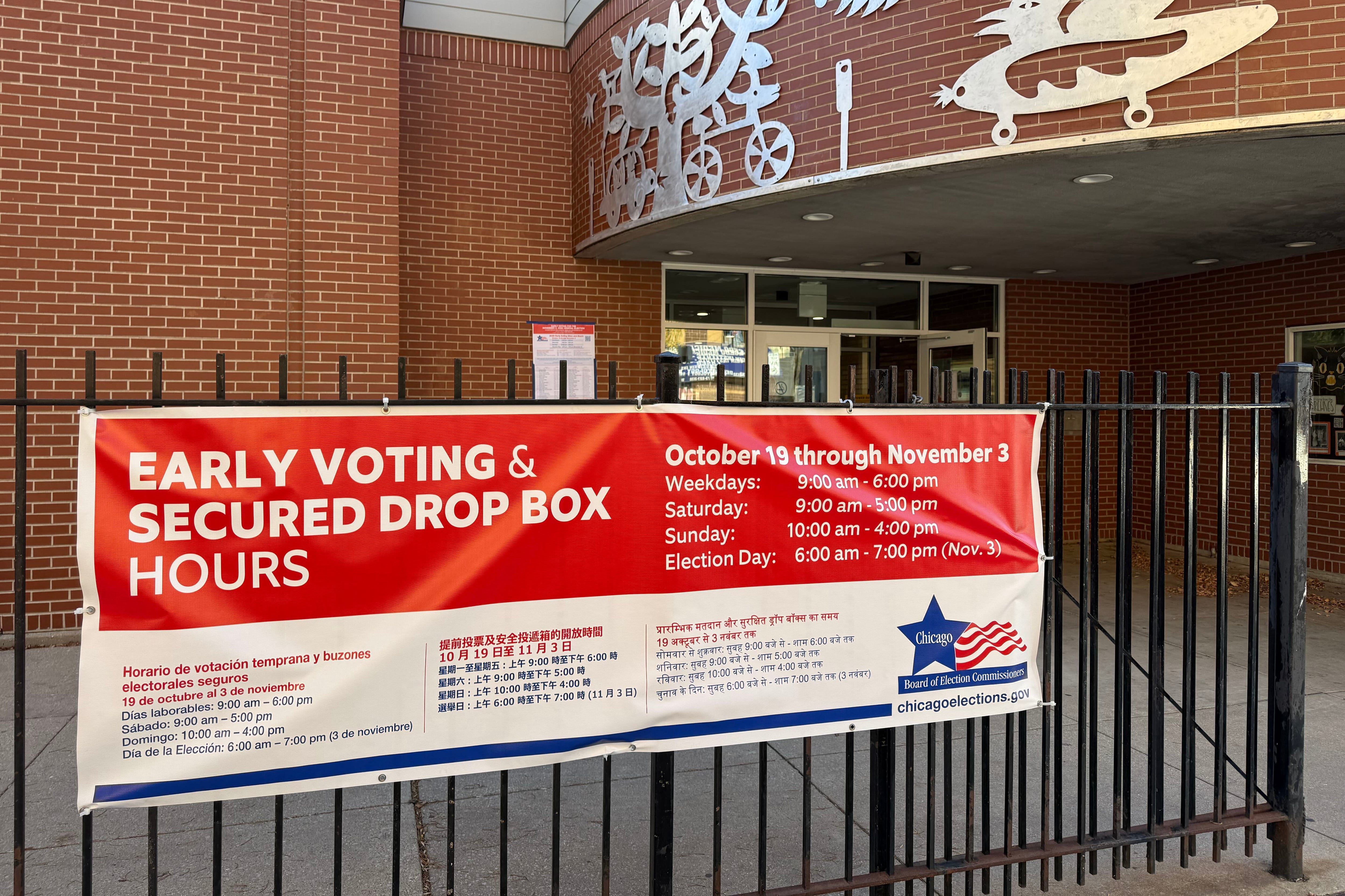 A large red and white sign hangs on a fence outside of a large brick building.