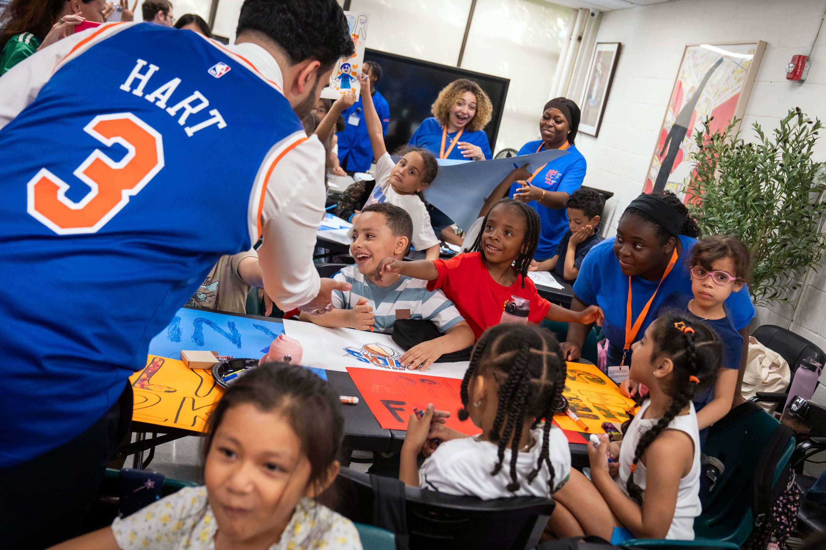 A man in a blue and orange Knicks jersey talks to kids who are making colorful posters.