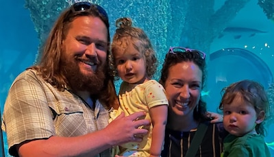 Two adults smile and hold two young children in front of an aquarium.