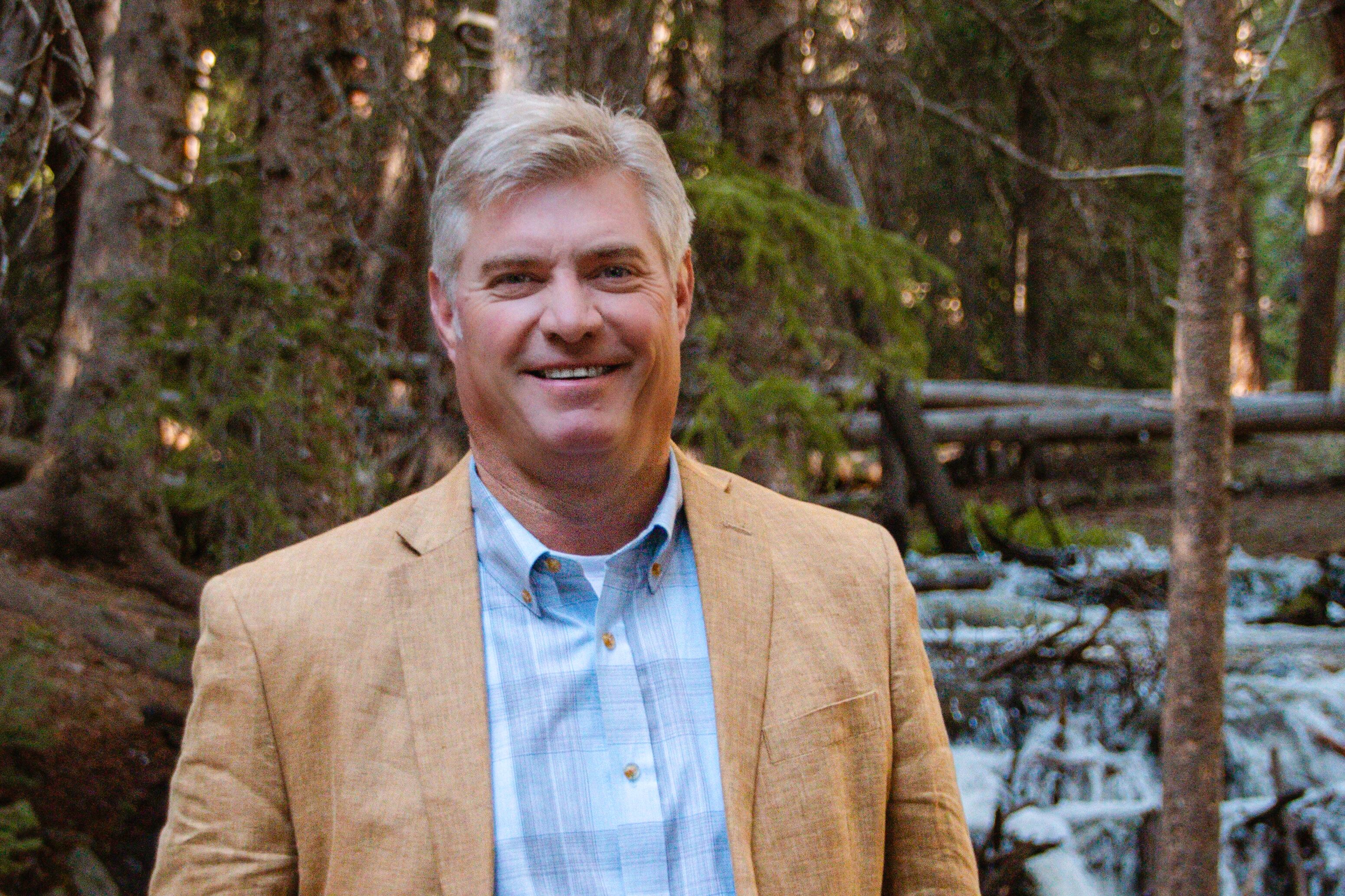 A portrait of a man wearing a tan suit jacket and a blue shirt standing outside with trees and a stream in the background.