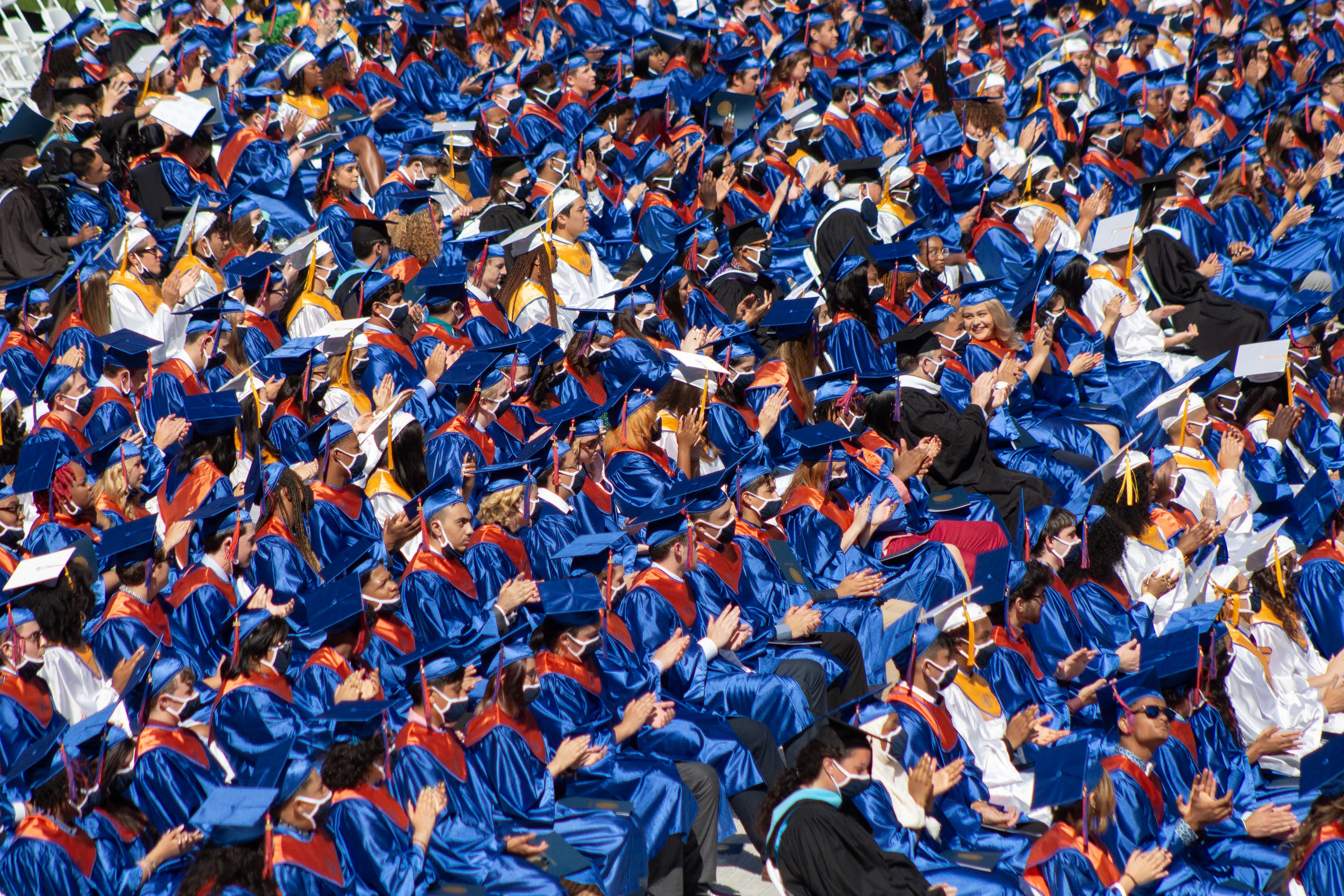 Rows of upcoming graduates applaud during a speech, creating a sea of blue and orange due to the shimmering of their caps and gowns from the sunlight.