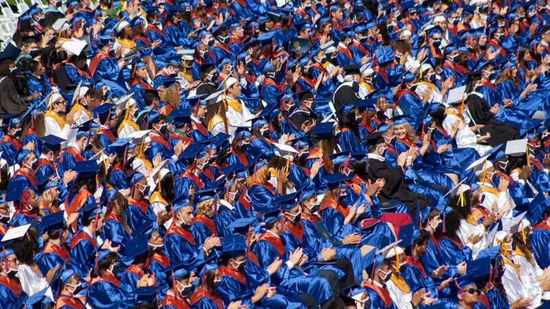Rows of upcoming graduates applaud during a speech, creating a sea of blue and orange due to the shimmering of their caps and gowns from the sunlight.