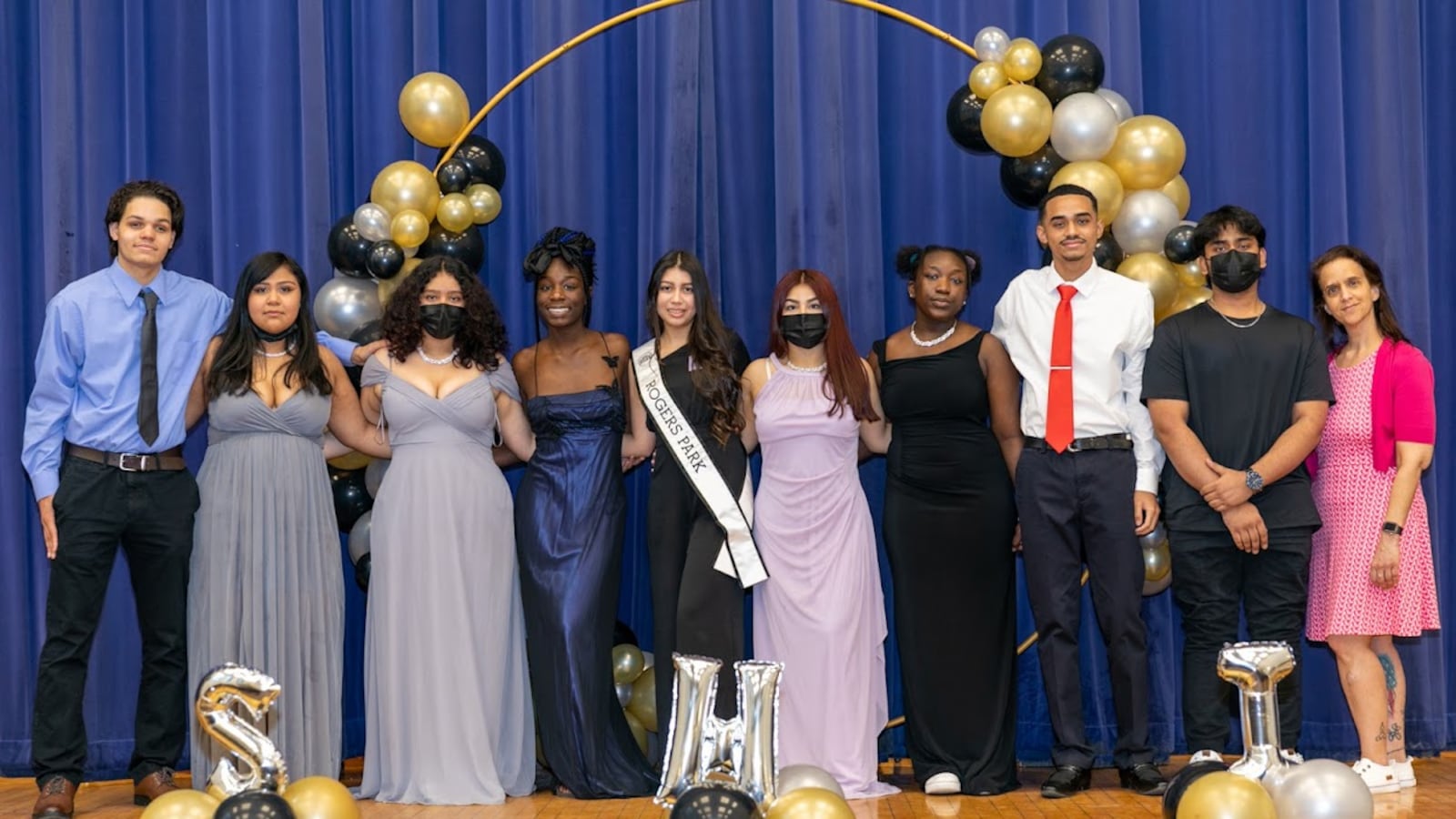 Students in prom attire pose for a portrait together on a stage with a blue curtain in the background, surrounded by celebratory gold and black balloons.