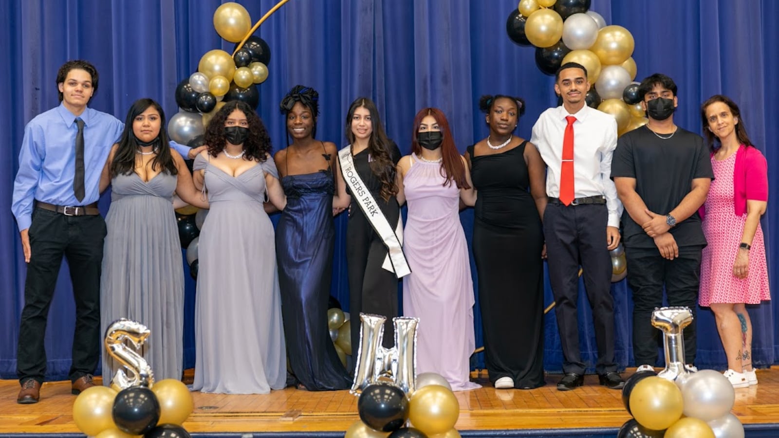 Students in prom attire pose for a portrait together on a stage with a blue curtain in the background, surrounded by celebratory gold and black balloons.