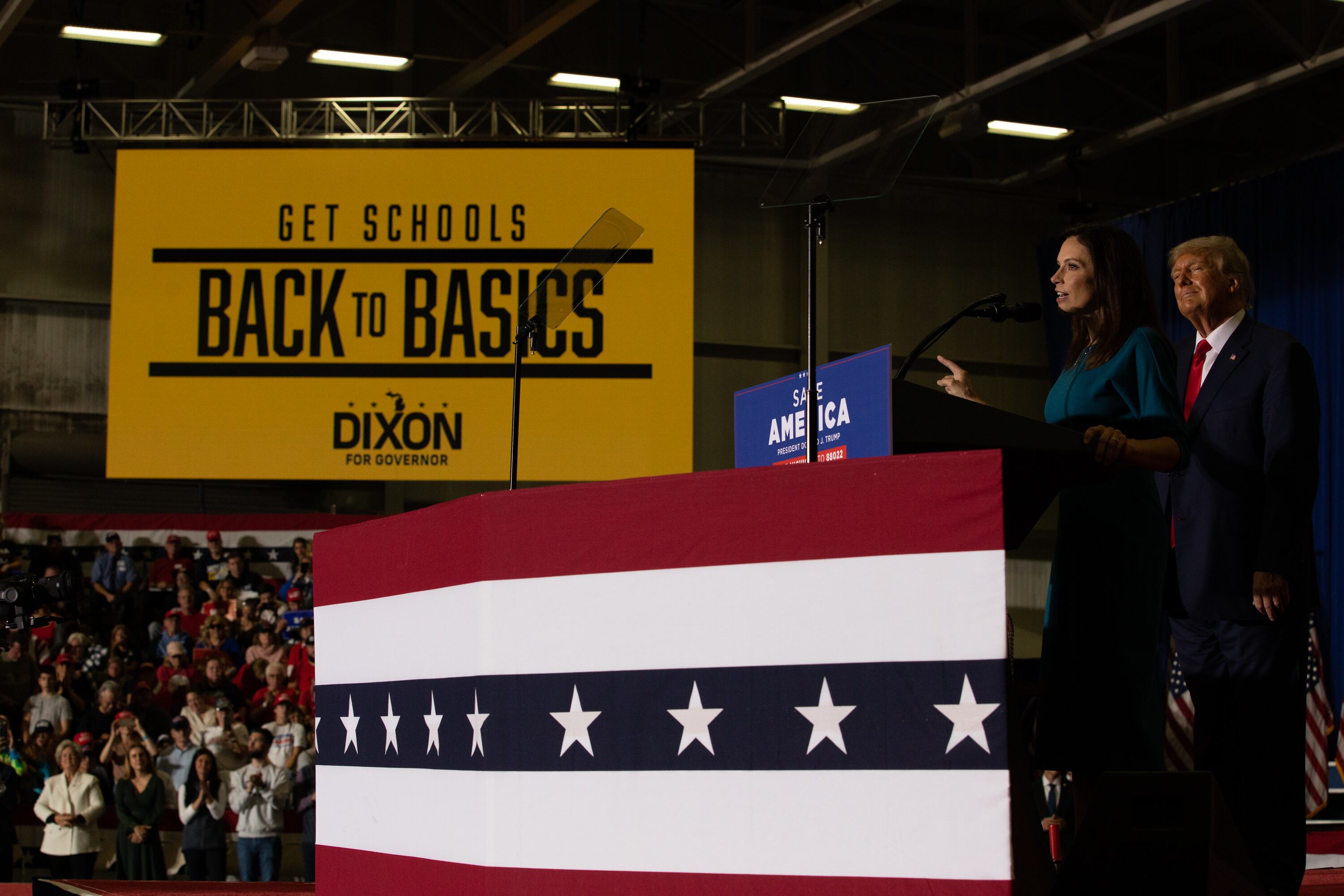 Tudor Dixon speaks at a microphone with Donald Trump standing behind her on a red-white-and-blue platform. A large yellow banner in the background says “Get Schools Back to Basics.”