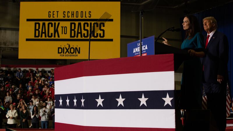 Tudor Dixon speaks at a microphone with Donald Trump standing behind her on a red-white-and-blue platform. A large yellow banner in the background says “Get Schools Back to Basics.” 