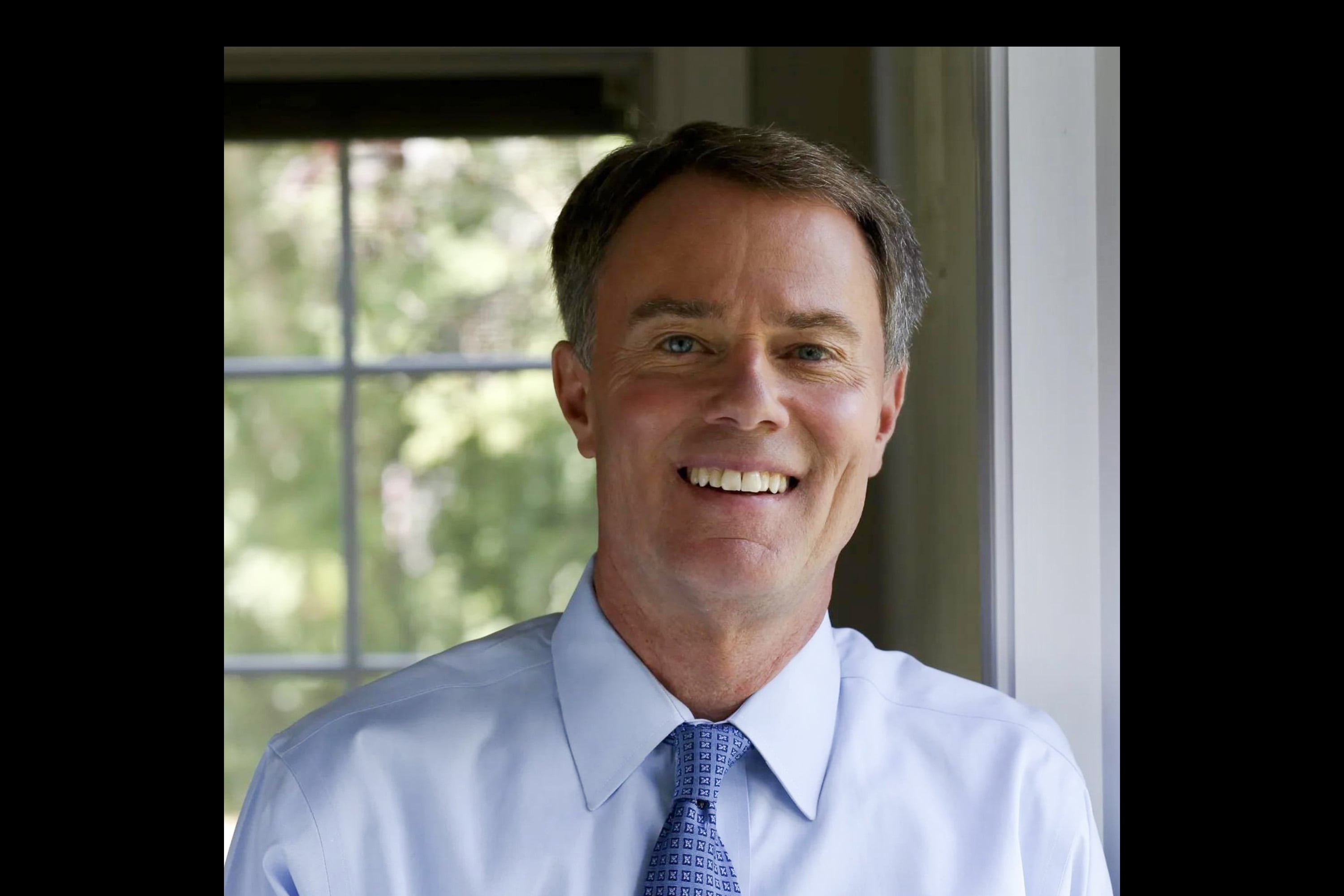 A man with light brown hair smiles for a portrait while wearing a white dress shirt and a blue tie in front of a window.