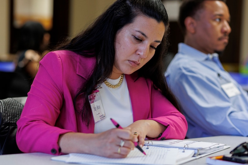 A person wearing a pink suit takes notes while sitting next to another person at a desk.