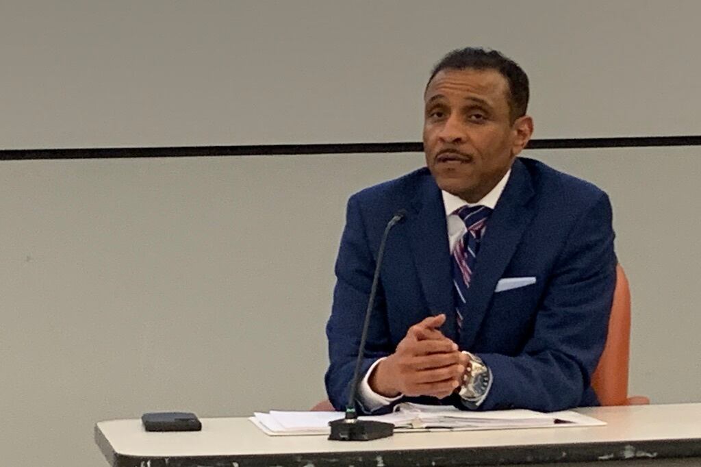 A man, wearing a blue suit with a blue and purple striped tie, speaks into a microphone during a town hall.