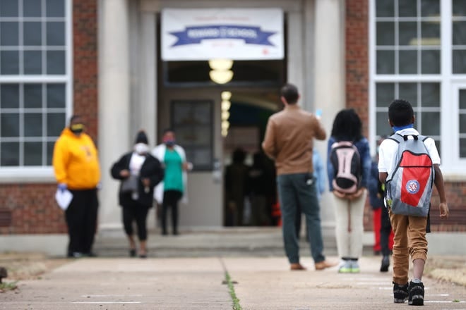 A young boy wearing a white shirt, khakis and a red and grey Shelby County Schools backpack walks into a school’s front doors.