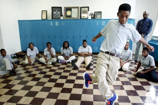 Eighth-grade boy in pale-blue shirt and beige trousers dances in front of classmates seated on a tile floor in front of lockers.