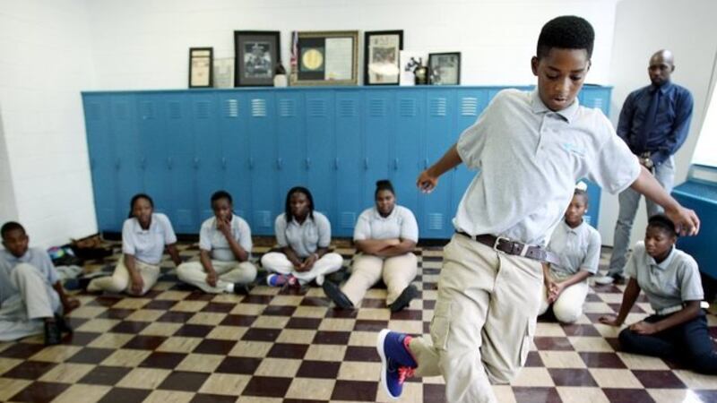 Eighth-grade boy in pale-blue shirt and beige trousers dances in front of classmates seated on a tile floor in front of lockers.