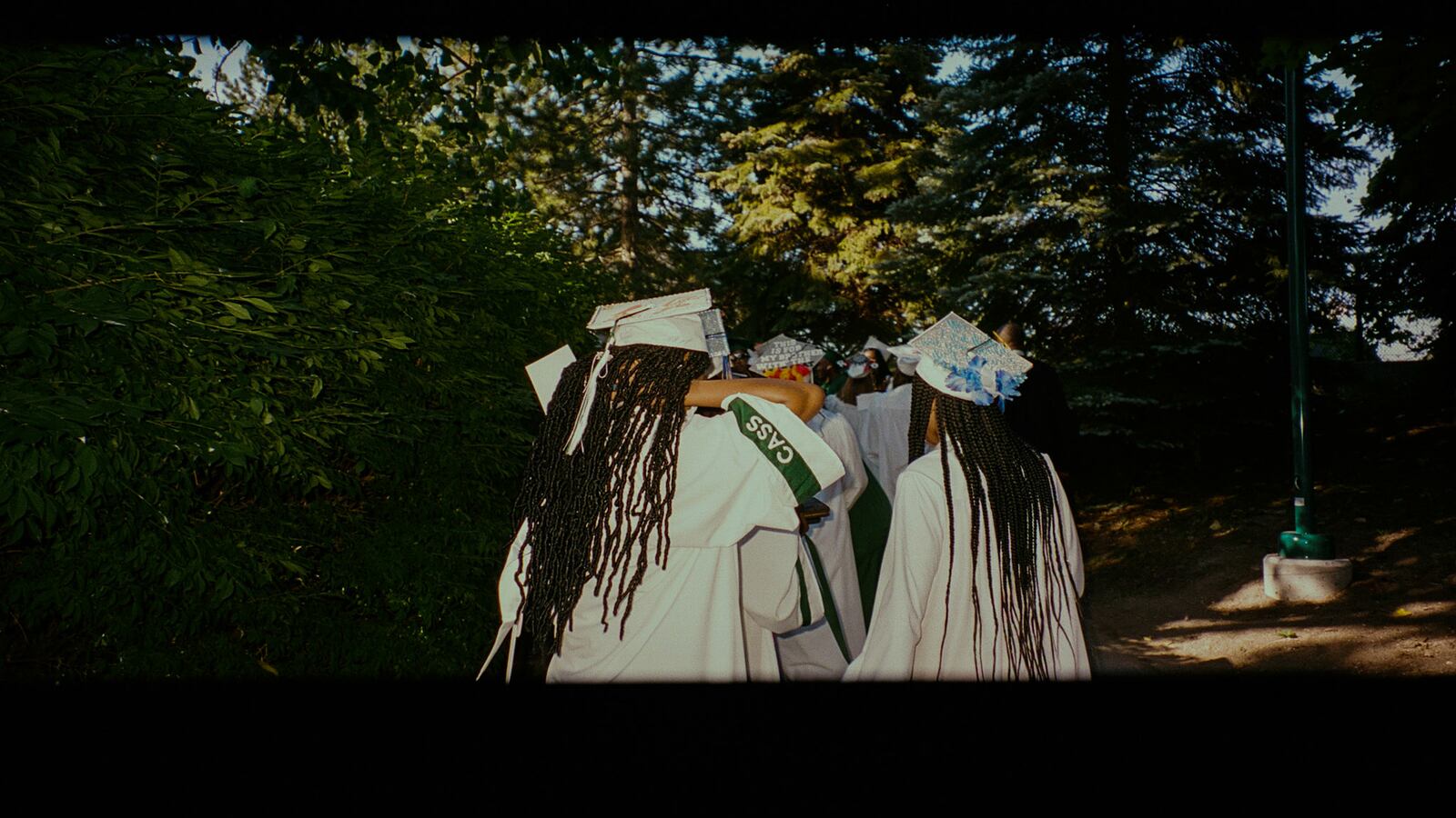 A group of young women wearing white graduation regalia make their way to their ceremony, their long braided hair running down their gowns with a green ribbon reading “CASS” on one of their arms.
