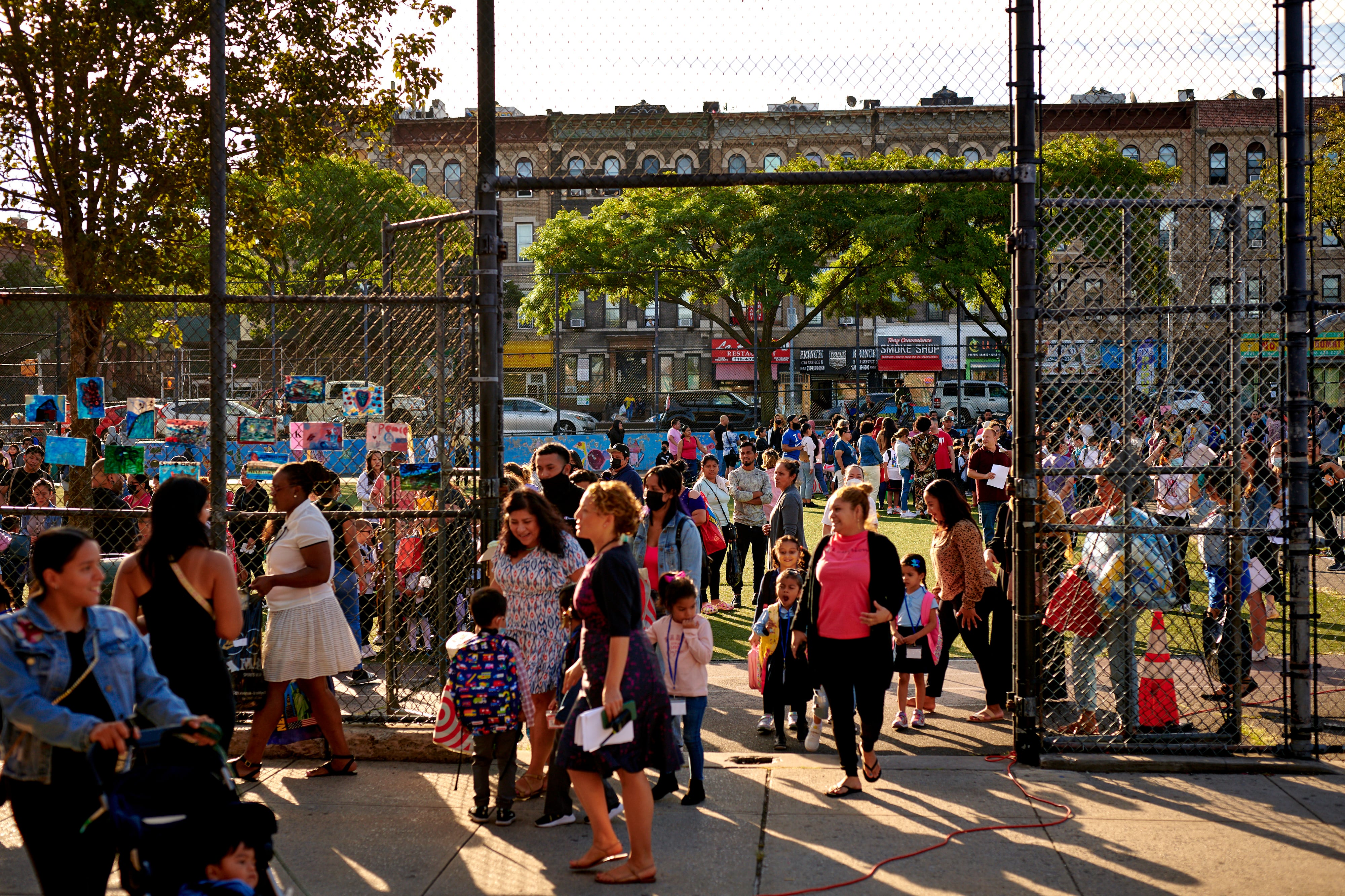 A large crowd of students and families stand outside of a school with a large wire fence.