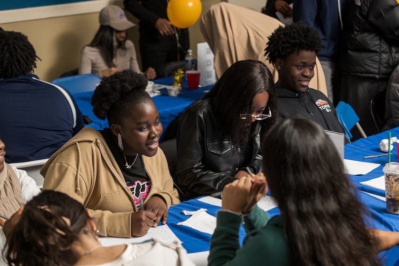 A group of high school students sit at tables with blue table cloths in a room.