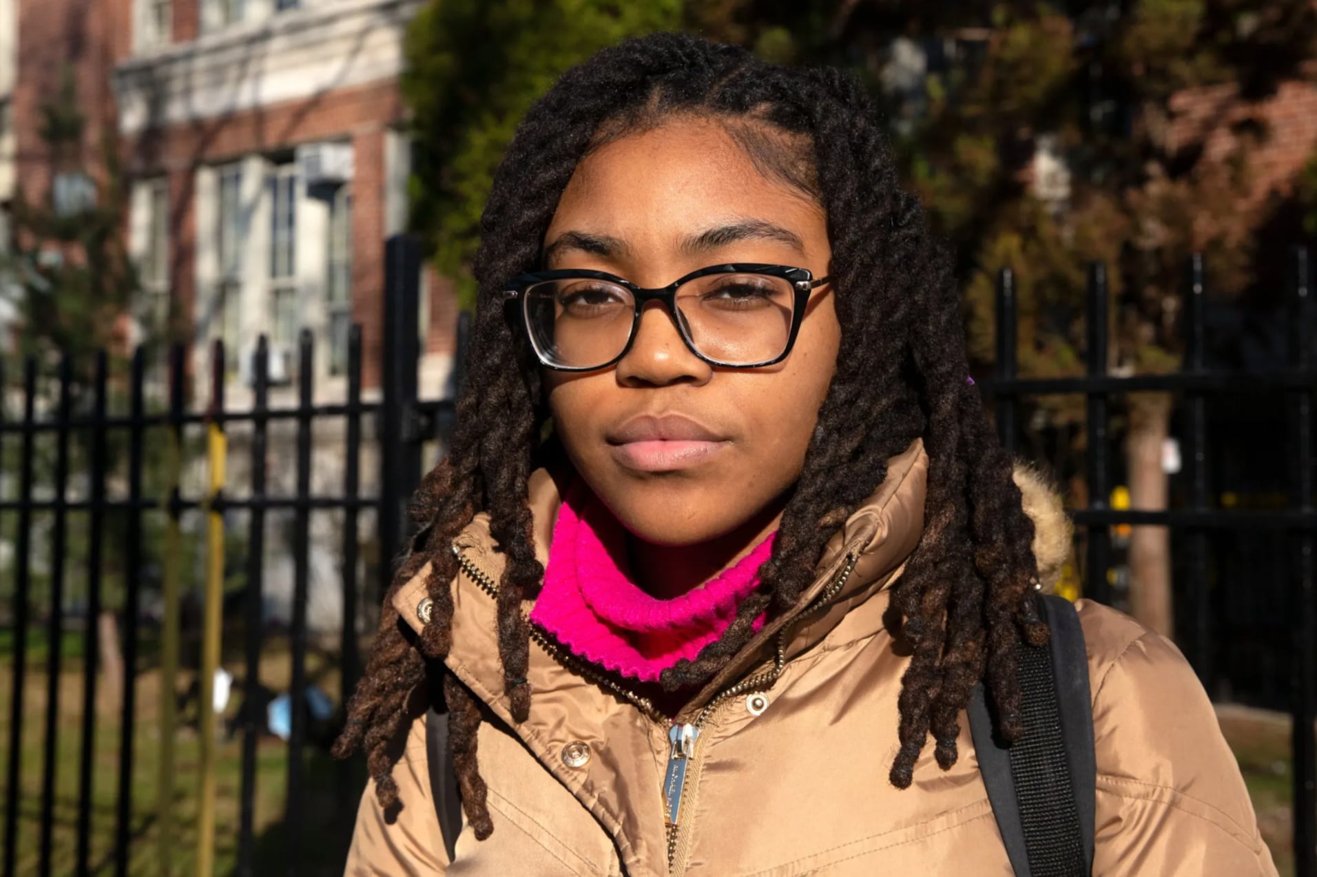 A girl with glasses, a pink scarf, and tan coat looks at the camera.
