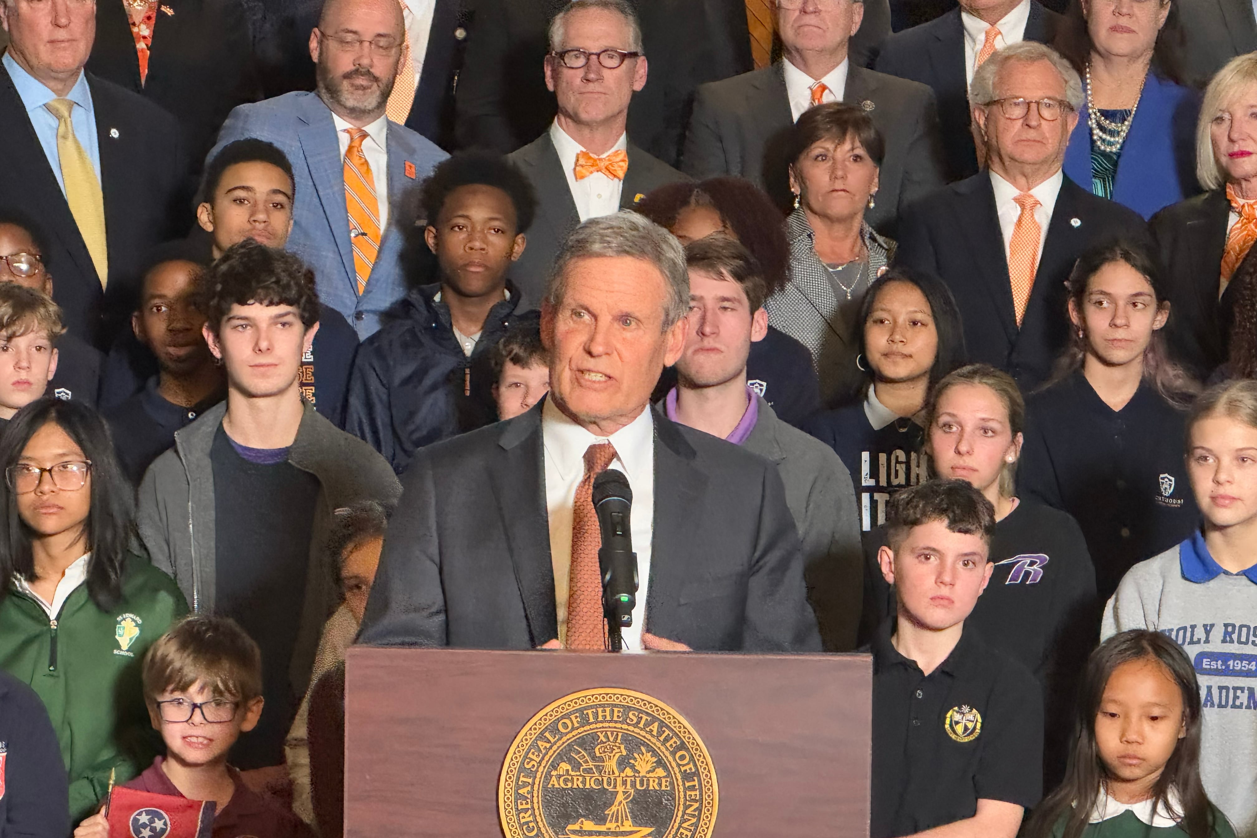 A man in a suit speaks from behind a podium with a group of children and adults in the background.