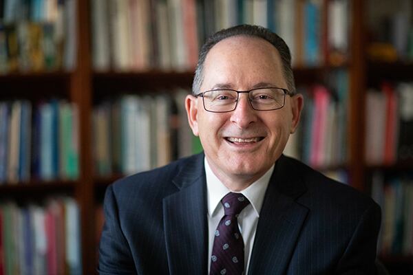 A man wearing a suit smiles at the camera for a portrait in front of a bookshelf full of books.