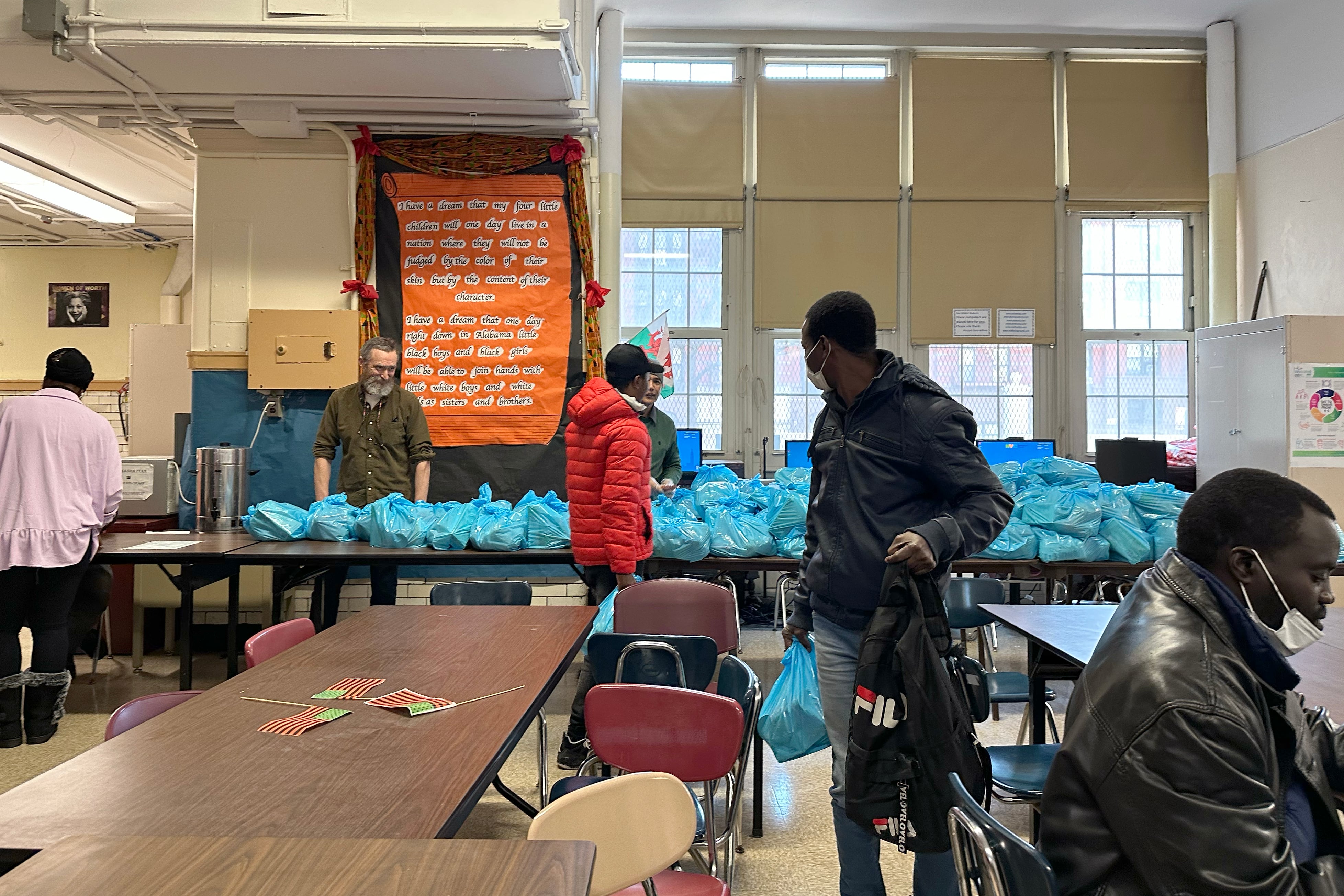 Blue bags line a long table in the background, while a few people stand in the foreground.