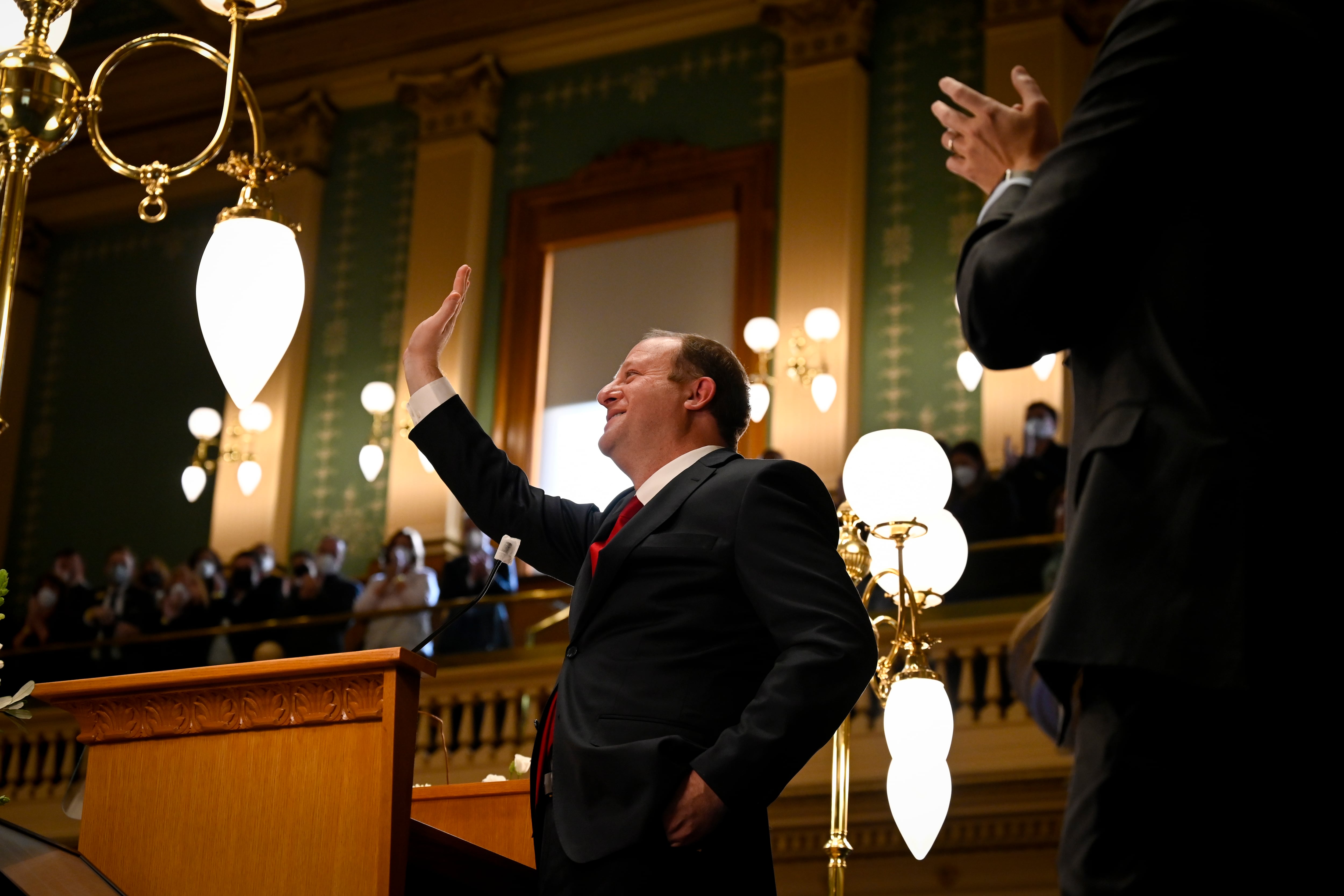 Colorado Gov. Jared Polis dressed in a black suit and red tie waves from the Colorado House chambers with lights and the green walls in the background.