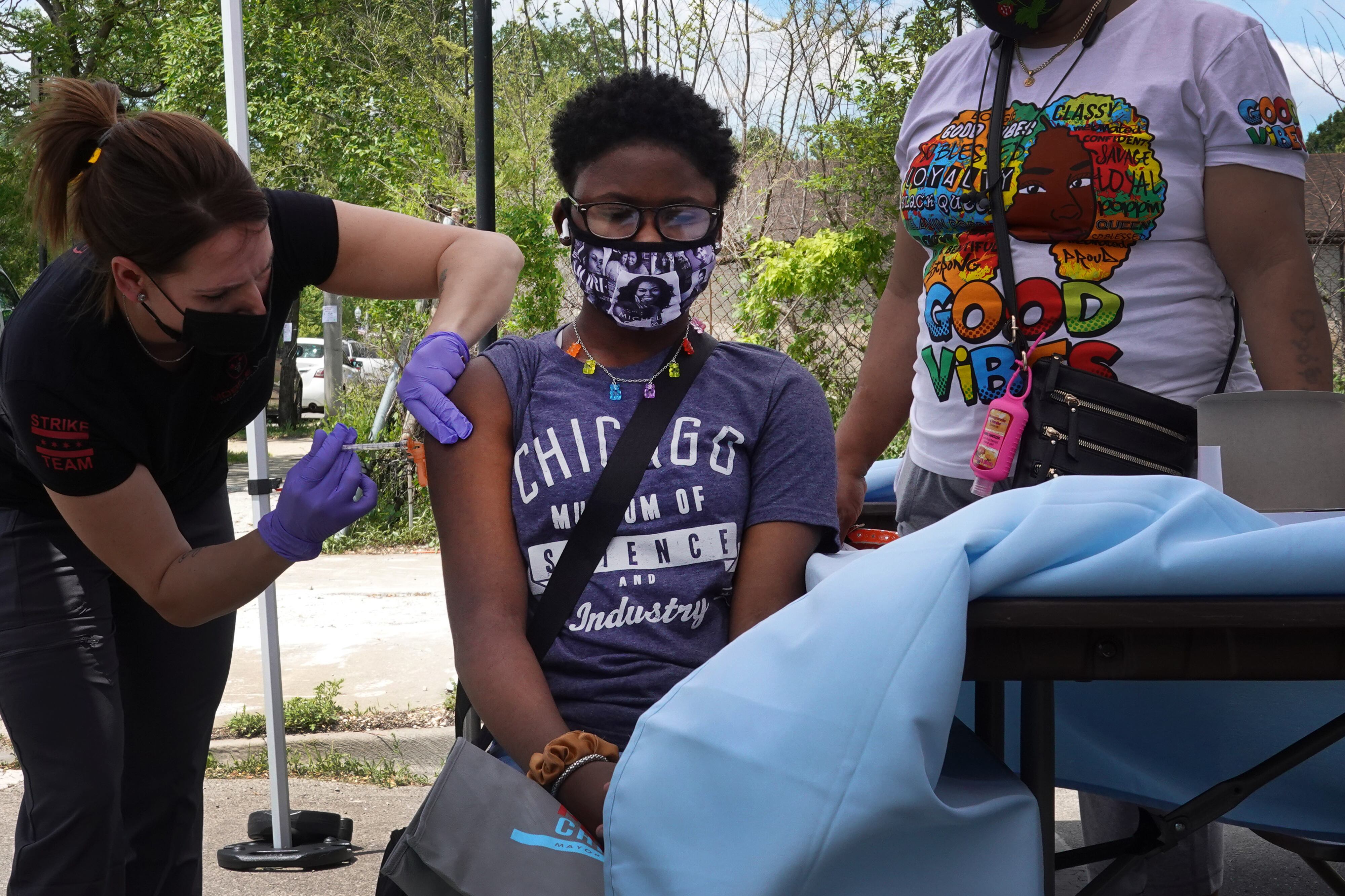 A girl wearing a Chicago shirt stands between two women, one of whom is giving the girl a dose of the COVID vaccine.