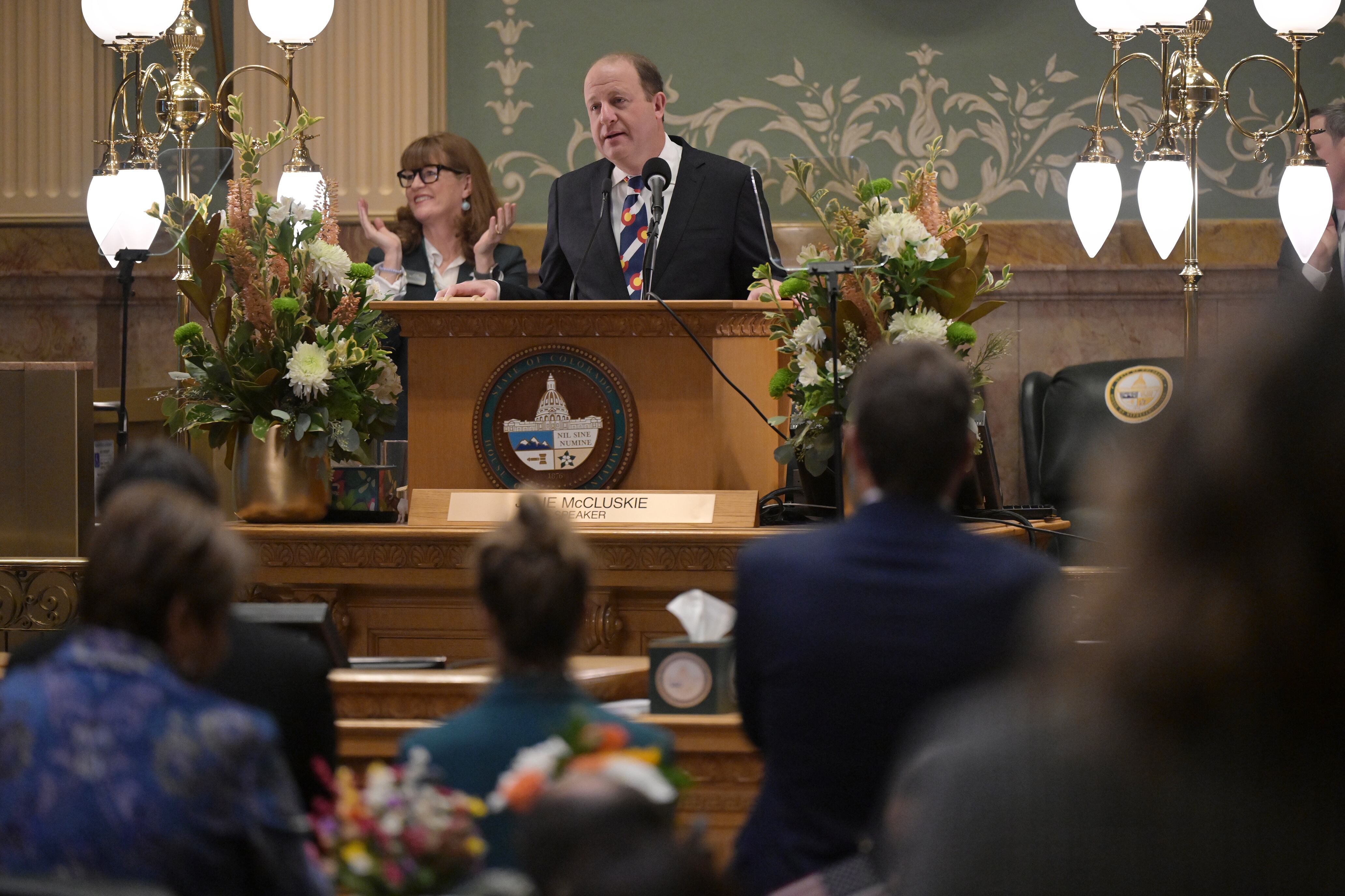 A man wearing a dark suit stands at a podium with a woman standing behind him and the back of people sitting in the foreground.