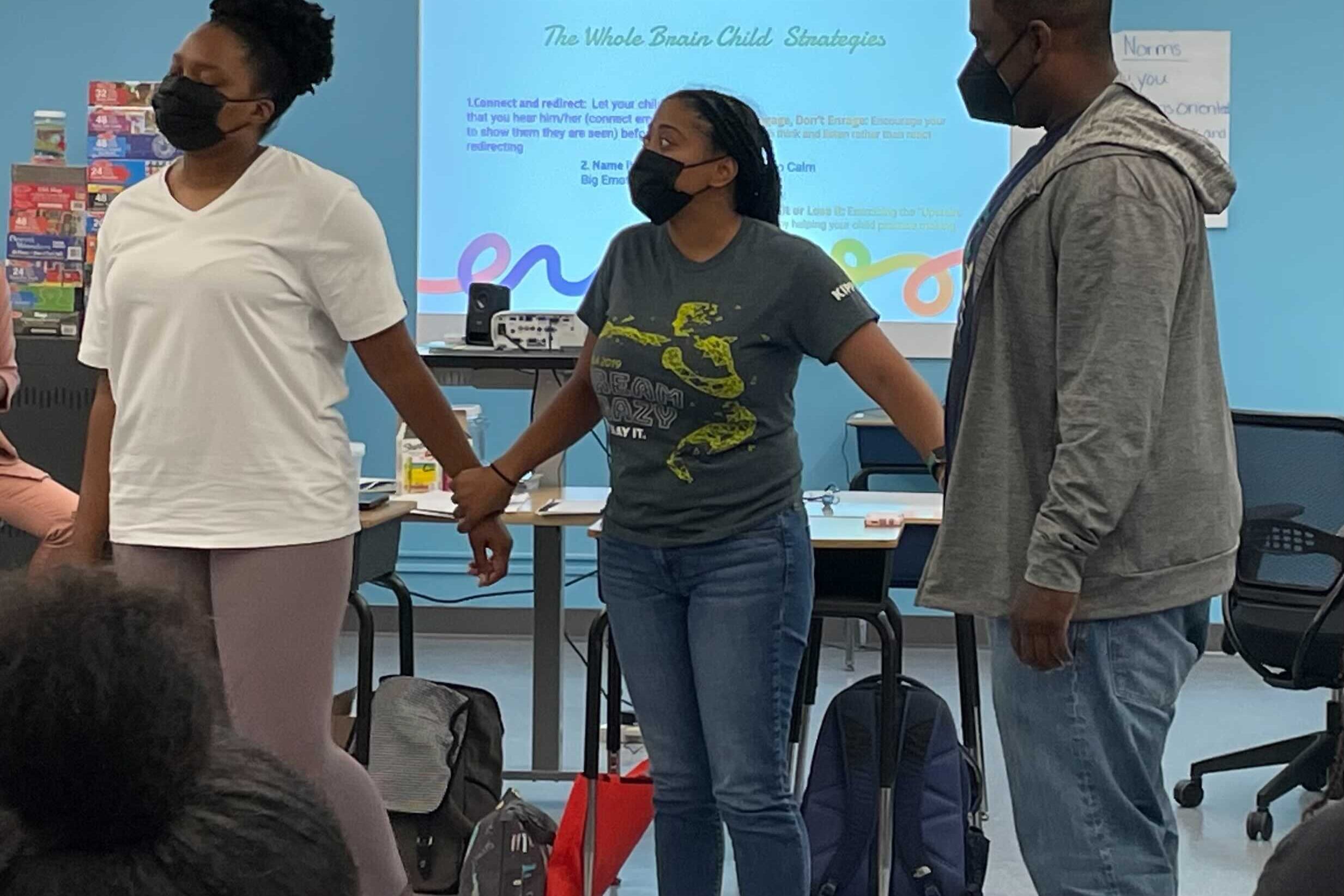 Three teachers, all wearing masks, hold hands at the front of a classroom during an exercise.
