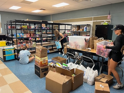 A photograph of three people working on various things in a large room with shelves and cardboard boxes with food and dry goods for students in need.