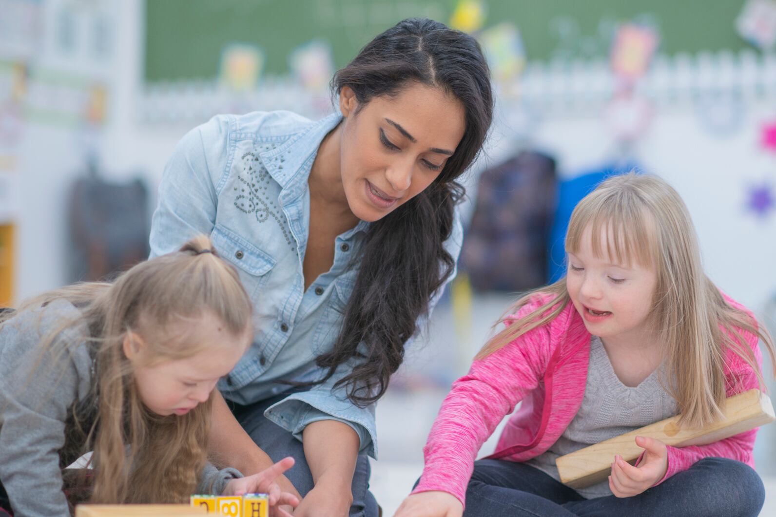 Two girls are sitting on the floor in an elementary school classroom. Their teacher sits in the middle while they play with wooden toy blocks.