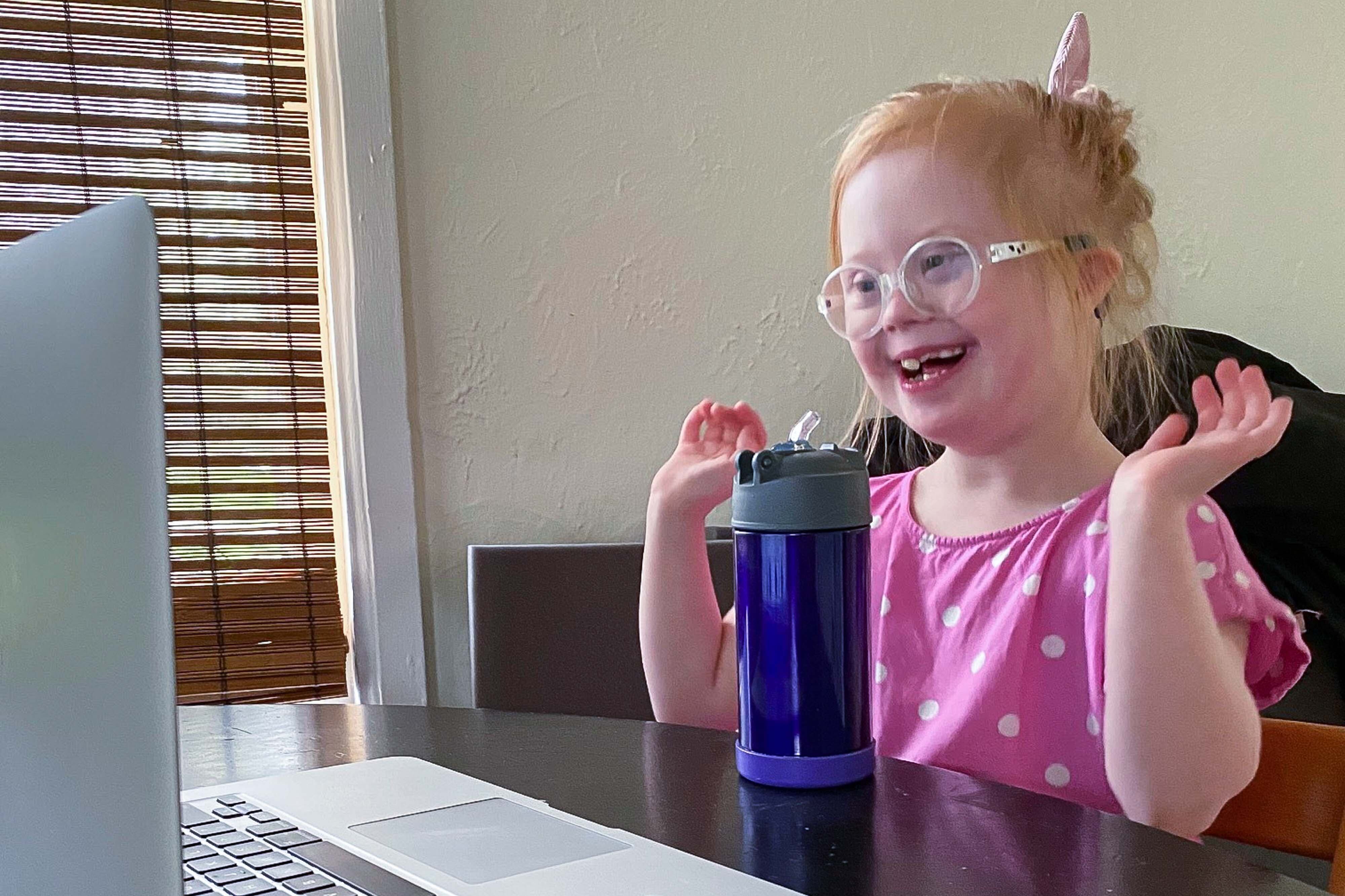 A little girl with Down syndrome smiles while looking at a computer screen. She has red hair and is wearing a pink polka-dotted shirt.
