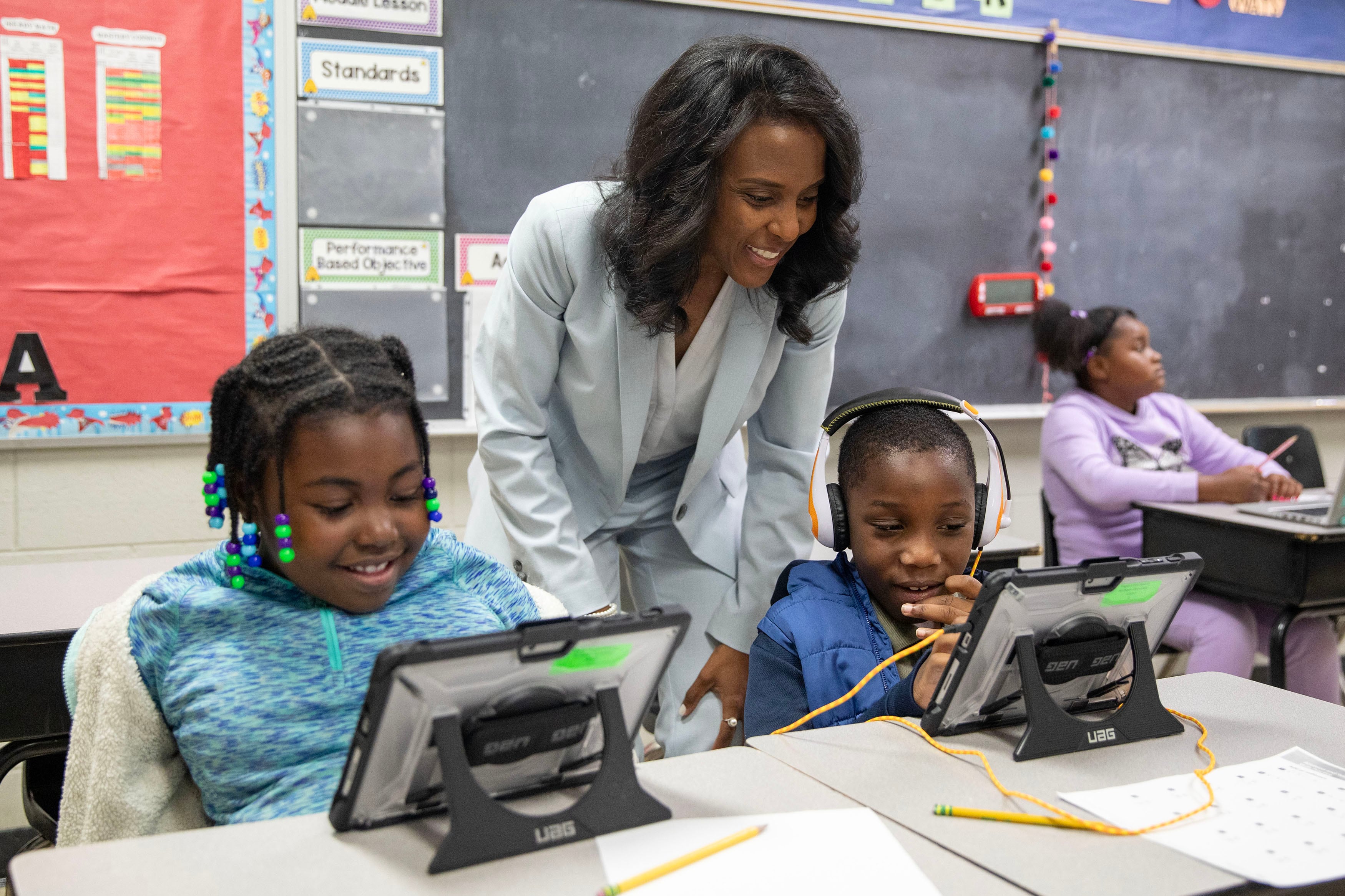 An adult in a blue suit stands between two students who are sitting at a desk wearing headphones and watching tablets.