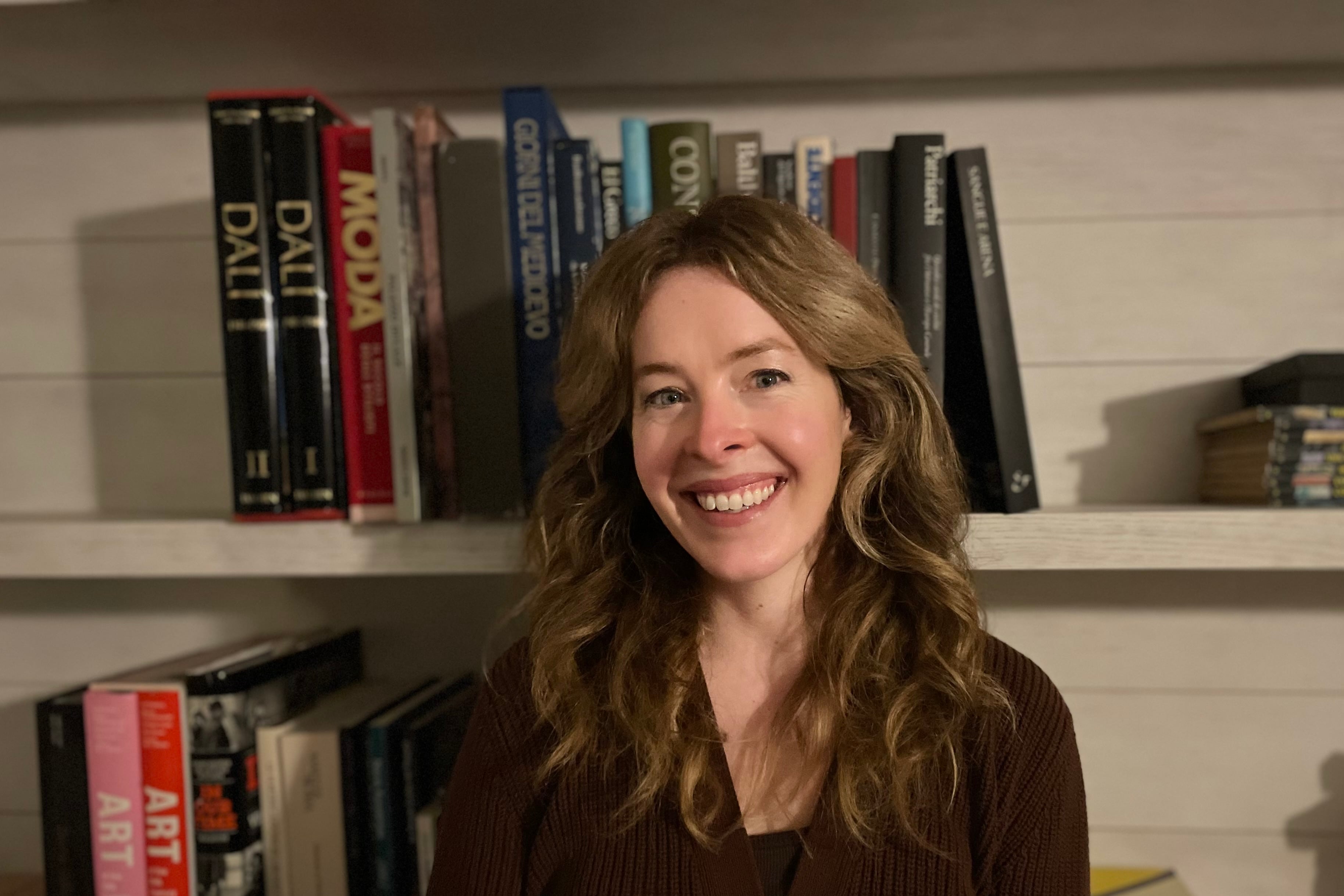 A woman wearing a dark sweater stands smiling in front of bookshelves.