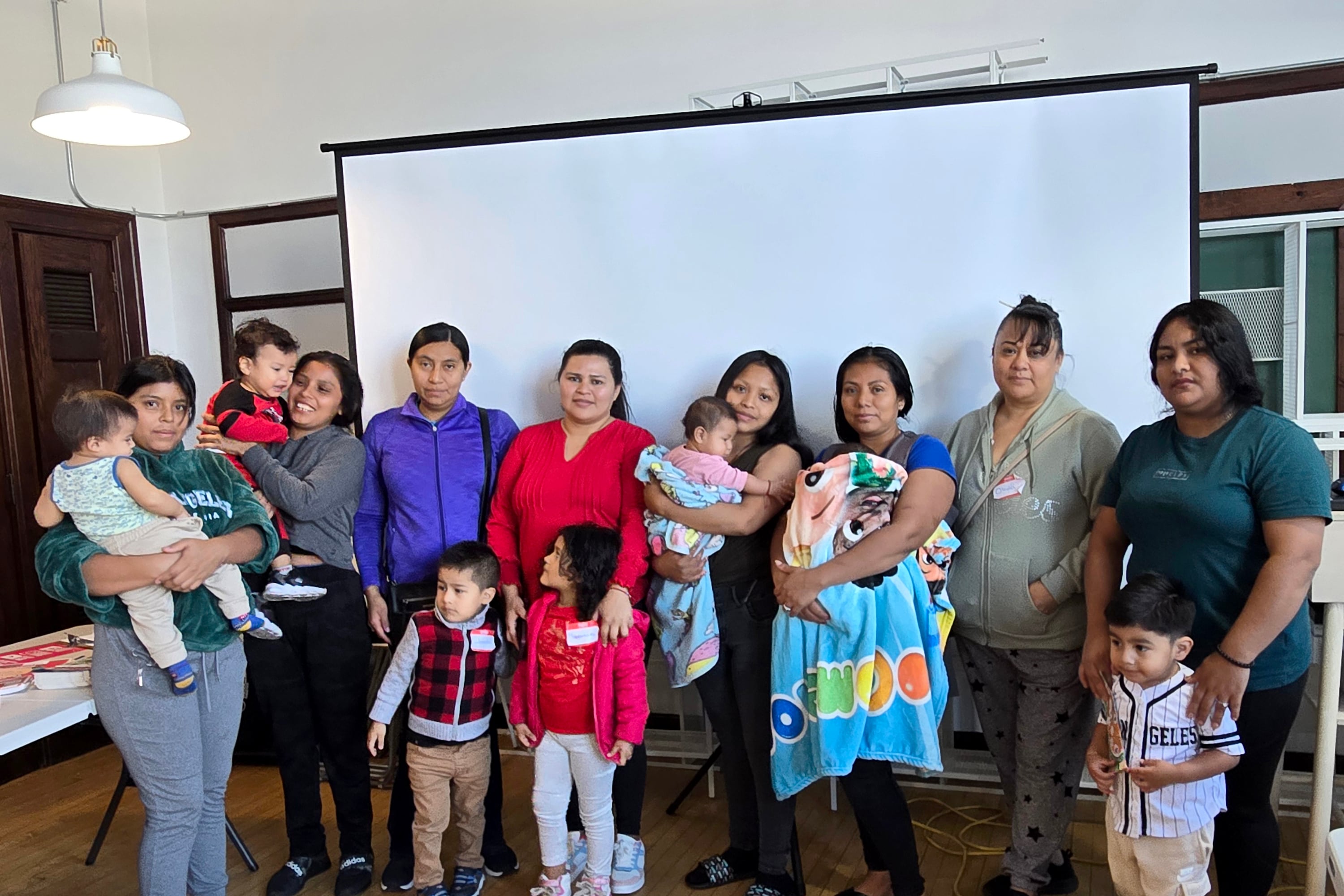 A group of Latine women, some holding young children, all pose for a photograph in front of a large screen in a room.