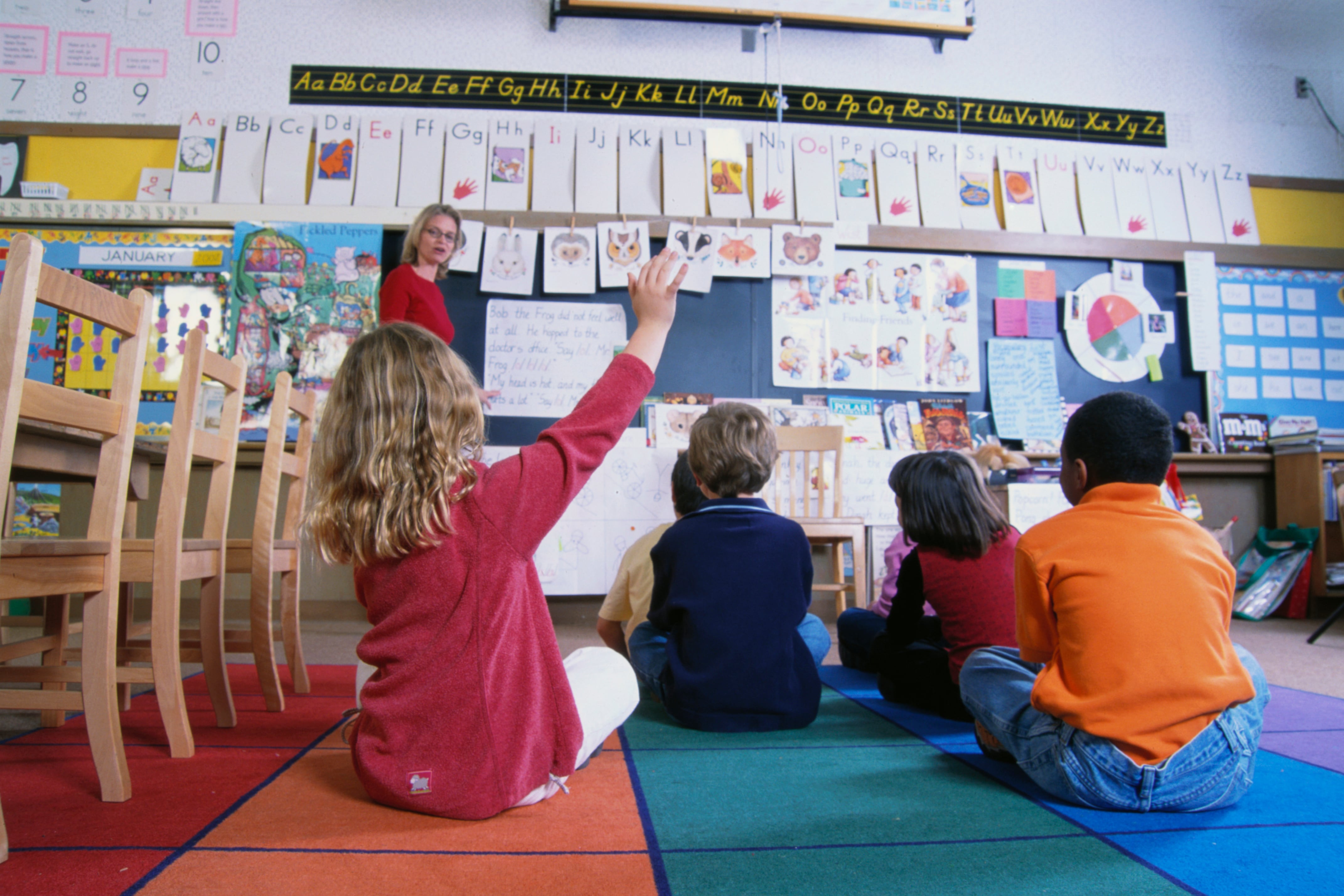 A group of students sits on the floor in a classroom with a teacher in front of them.