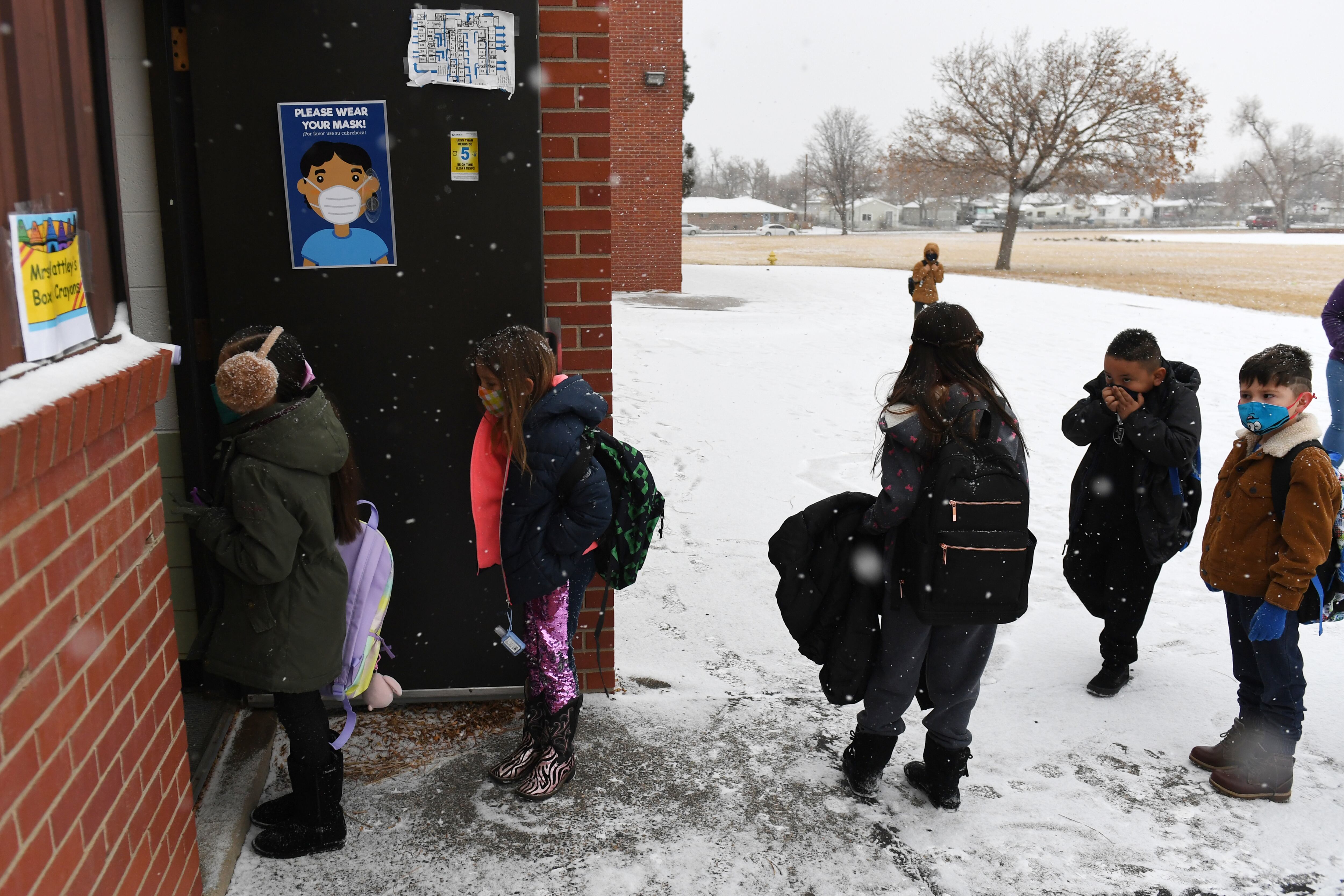 Young children in masks and winter coats line up to enter an Adams 14 school building on a snowy day.