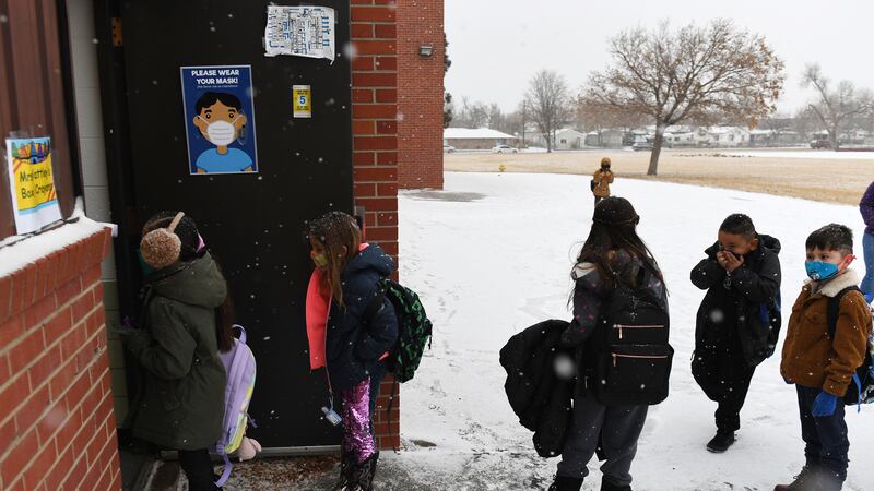 Young children in masks and winter coats line up to enter an Adams 14 school building on a snowy day.