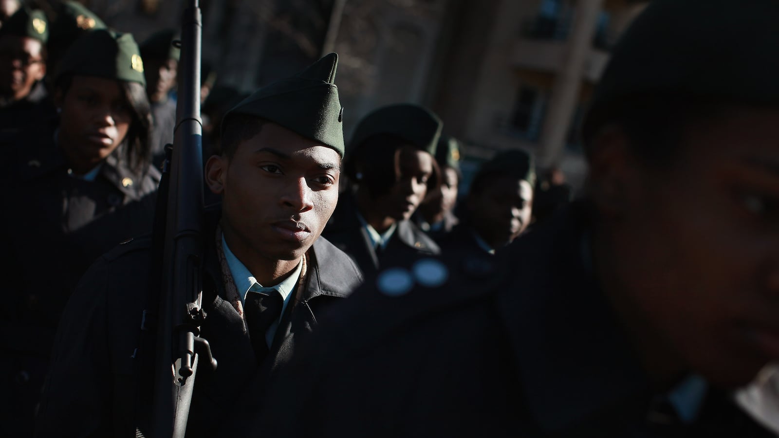 JROTC cadets, wearing full dress uniforms and holding wooden rifles, march in formation at a parade.