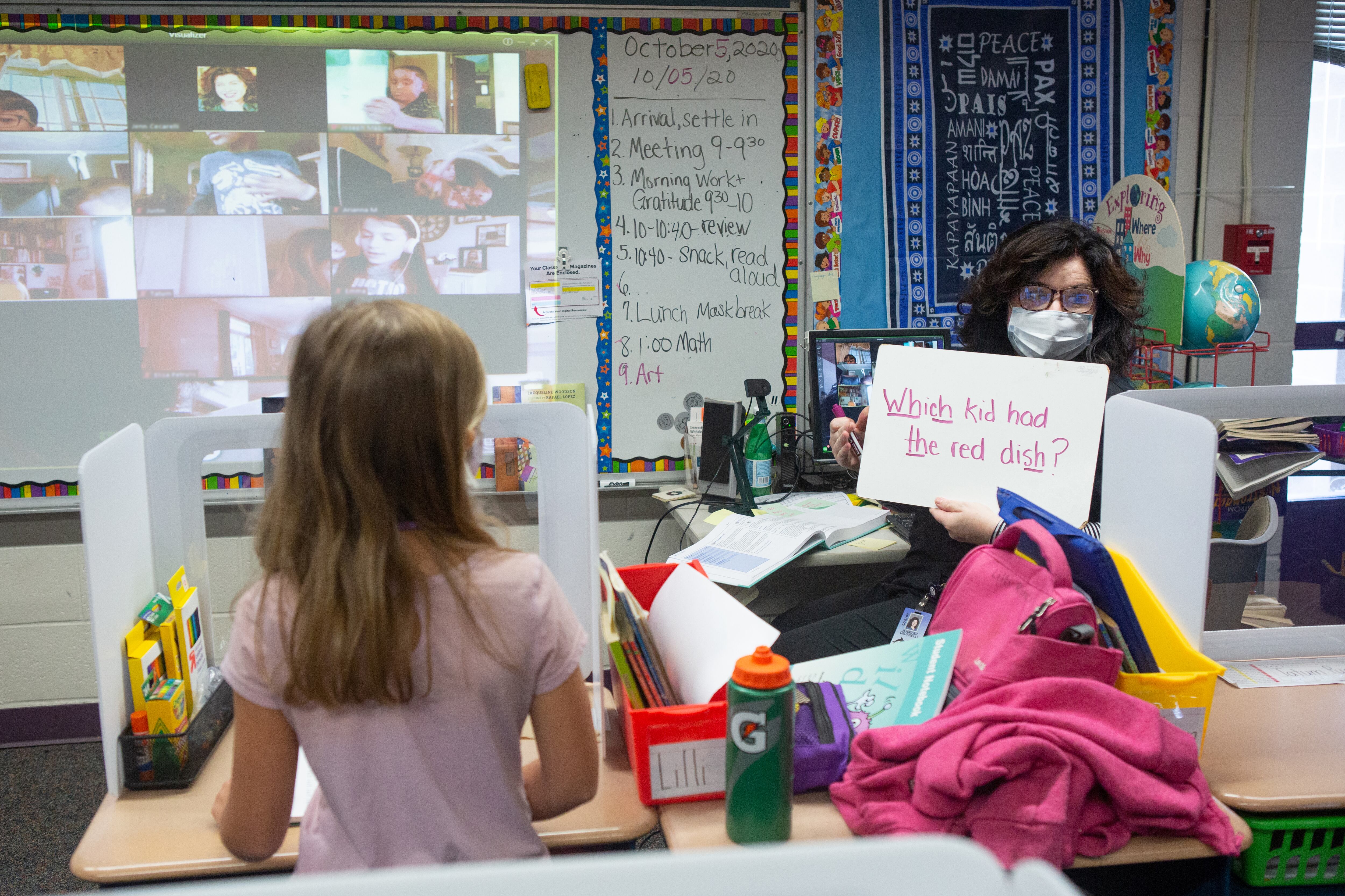 Second grade teacher Mrs. Cecarelli, wearing a mask, holds up a placard with the words, “Which kid had the red dish” with the digraphs Wh, ch, th and sh underlined. A girl in a pink top stands in the foreground in a classroom, while a board with a projection of students participating remotely is in the background, at Wesley Elementary School in Middletown, CT, October 5, 2020.