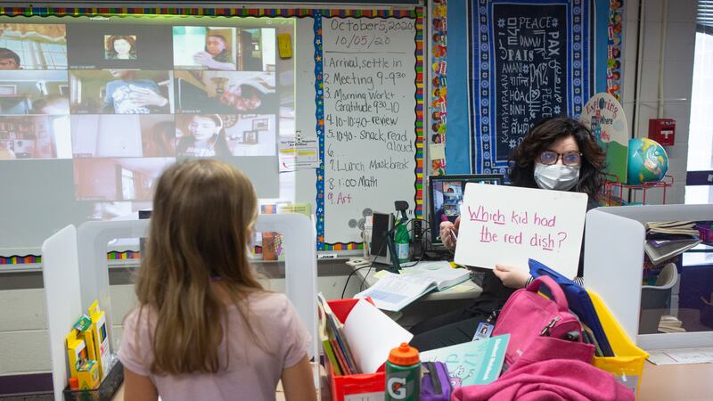 Second grade teacher Mrs. Cecarelli, wearing a mask, holds up a placard with the words, “Which kid had the red dish” with the digraphs Wh, ch, th and sh underlined. A girl in a pink top stands in the foreground in a classroom, while a board with a projection of students participating remotely is in the background, at Wesley Elementary School in Middletown, CT, October 5, 2020.