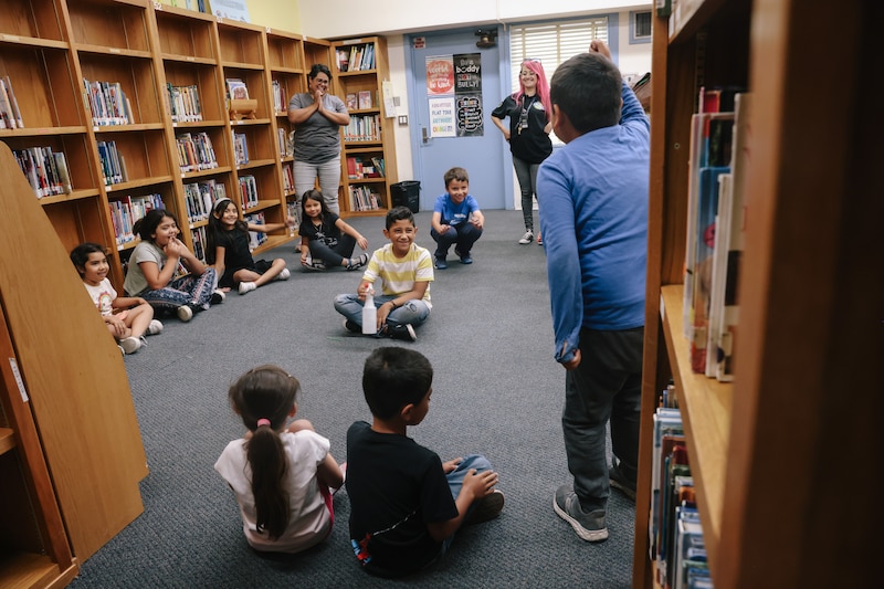 Students sit in a circle in a library with gray carpet.