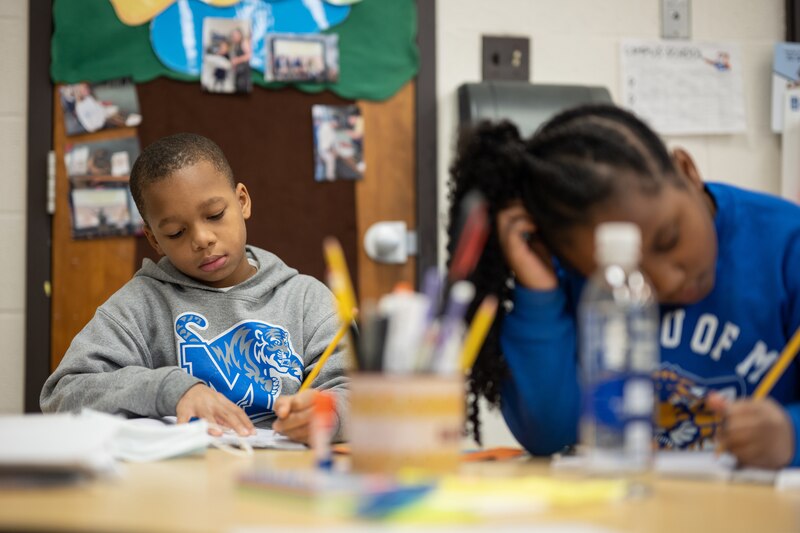 Two young students sit at their desks concentrating on their schoolwork while wearing blue, white and grey sweaters.